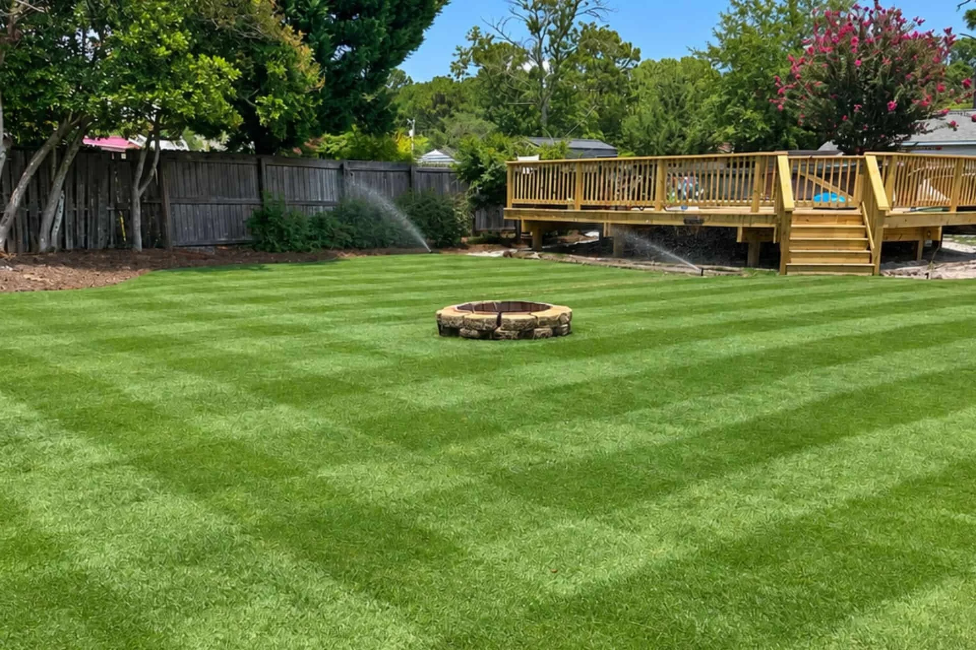 Striped, healthy backyard lawn with a fire pit and deck, irrigation system active, in Wilmington, NC