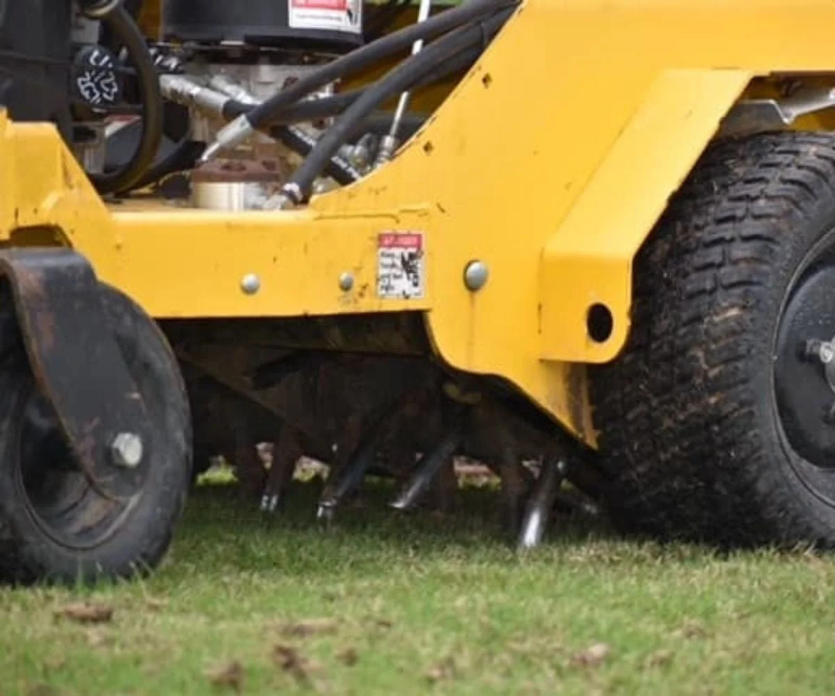 Close-up of a yellow lawn aerator on grass, showing wheels, blades, and hydraulic components.