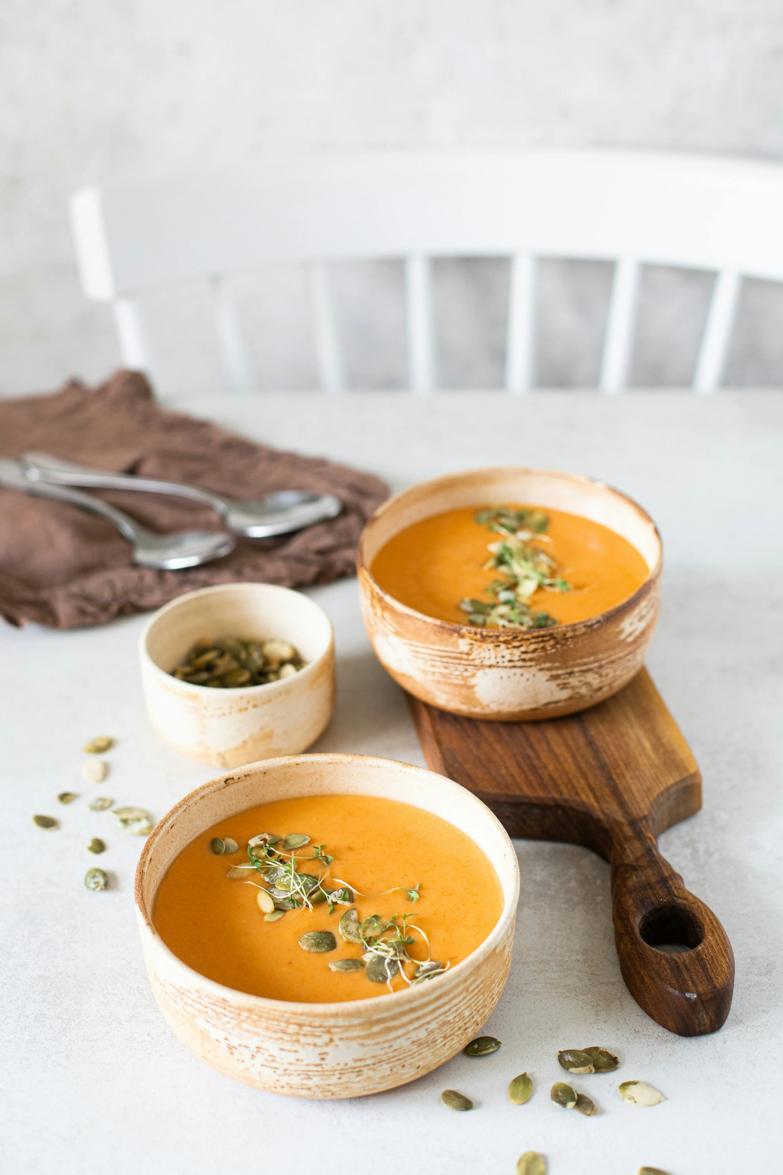 A white table features two bowls of orange-colored pumpkin soup garnished with microgreens and pumpkin seeds. A small bowl of pumpkin seeds and a wooden serving board with a soup bowl on it are also visible, along with a brown cloth napkin and two spoons.
