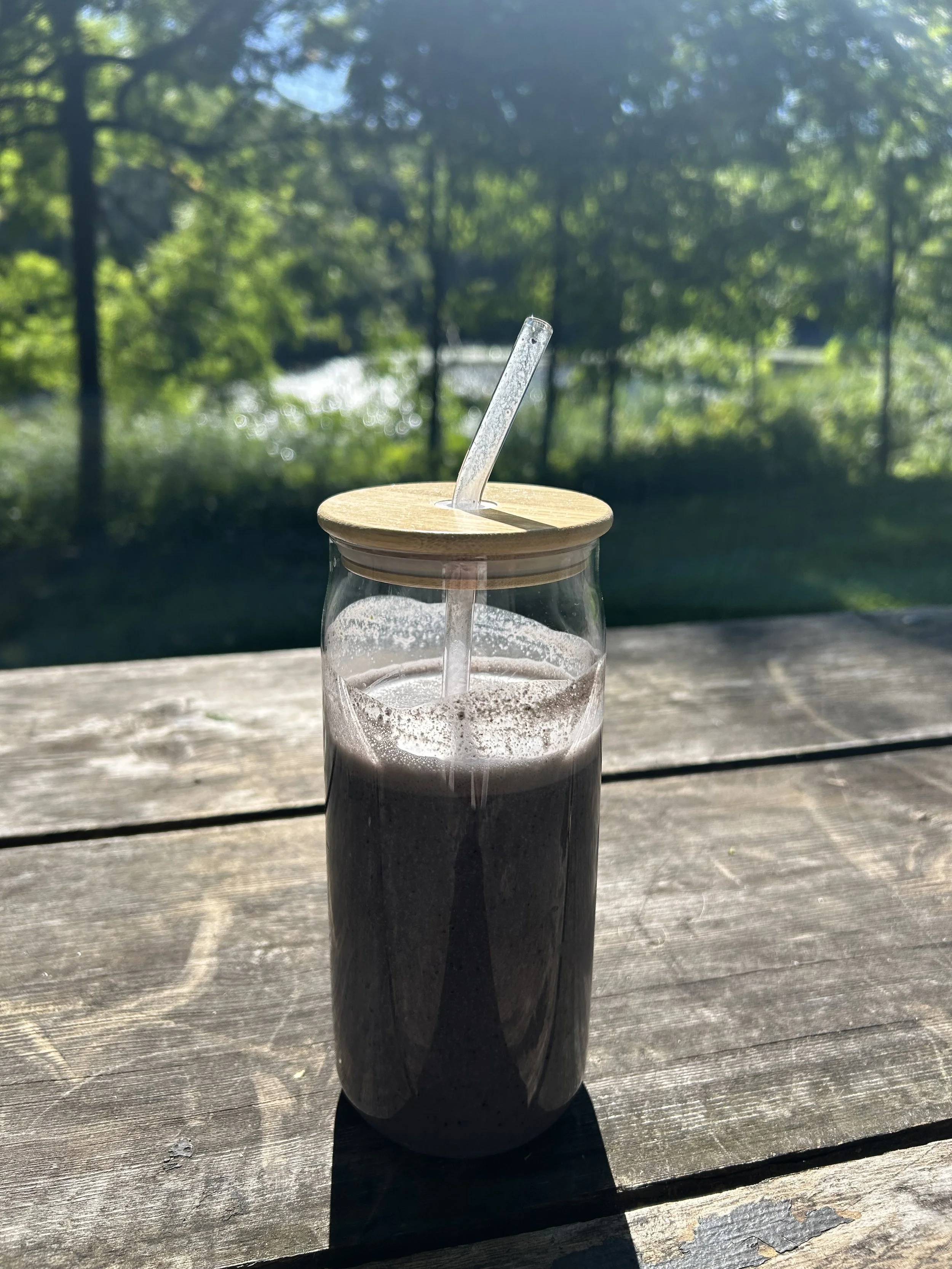 A glass bottle with a wooden lid and a straw, sitting on a wooden table outdoors with trees and sunlight in the background.