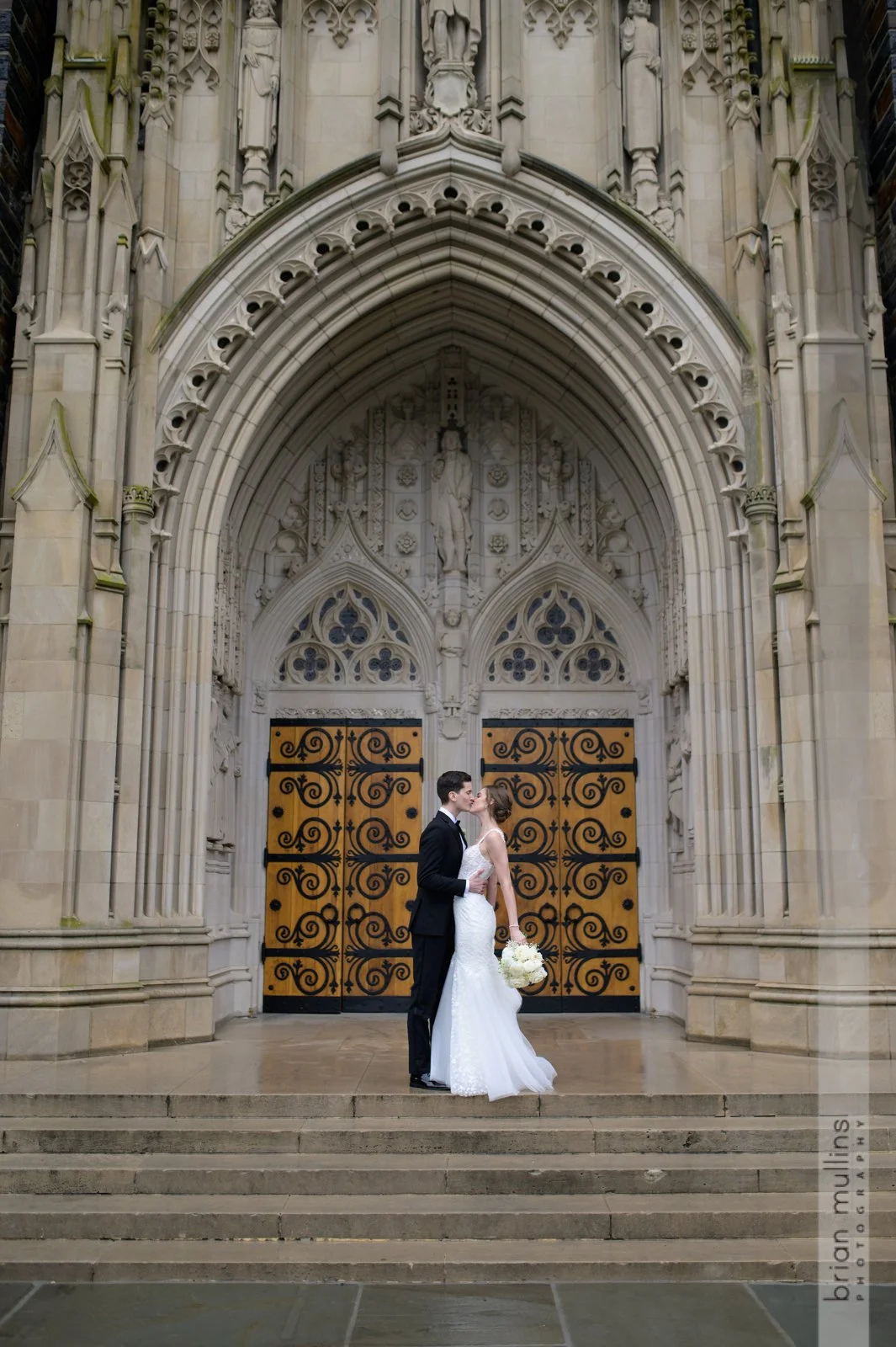A bride and groom share a kiss in front of a large Gothic-style church entrance with ornate stone carvings and wooden doors with decorative ironwork.