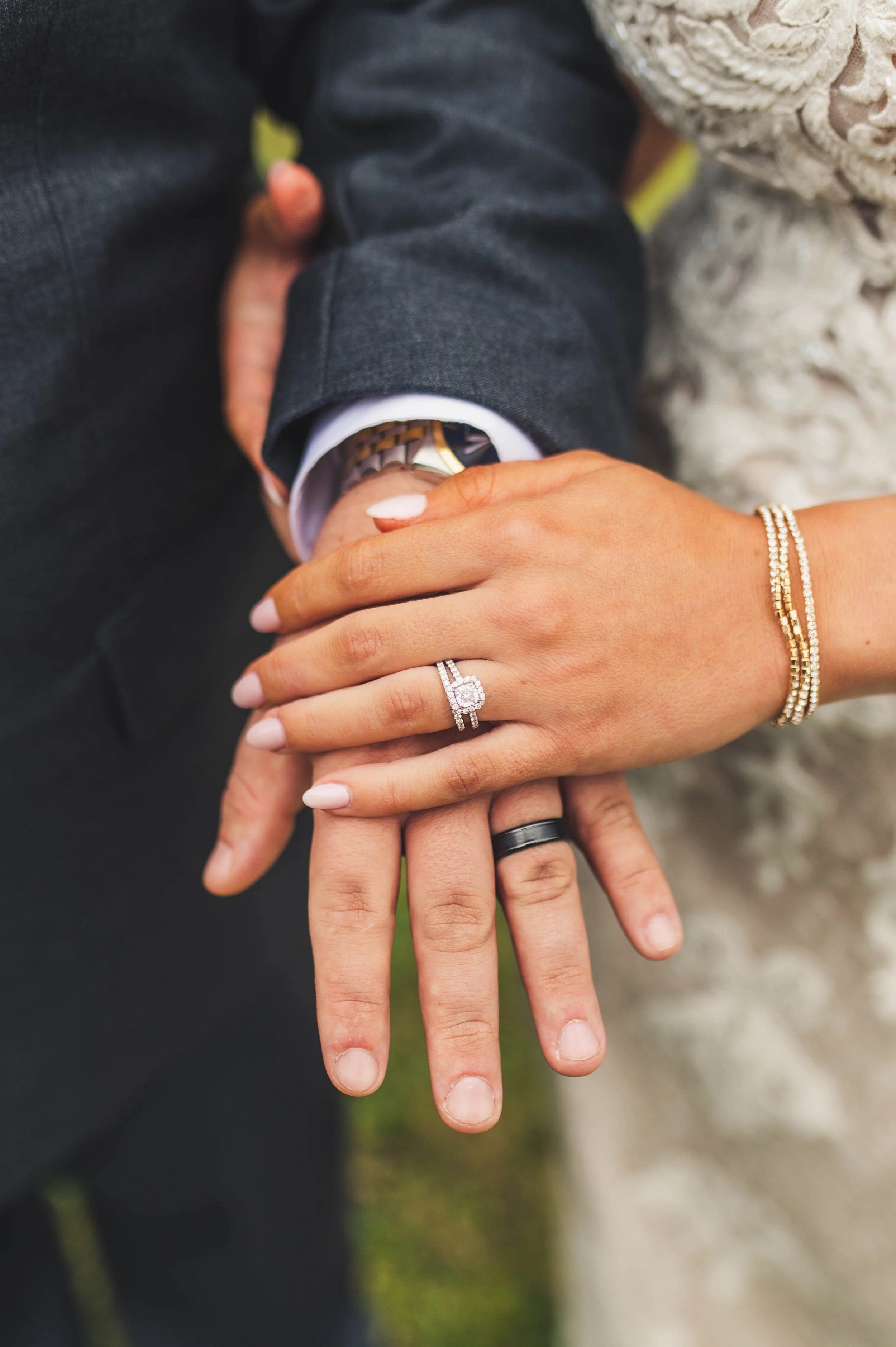 Close-up of a couple's hands showing wedding rings as they hold each other, with the man wearing a dark suit and the woman wearing a pearl bracelet and a ring with a large diamond. Their hands are intertwined, displaying their wedding bands.