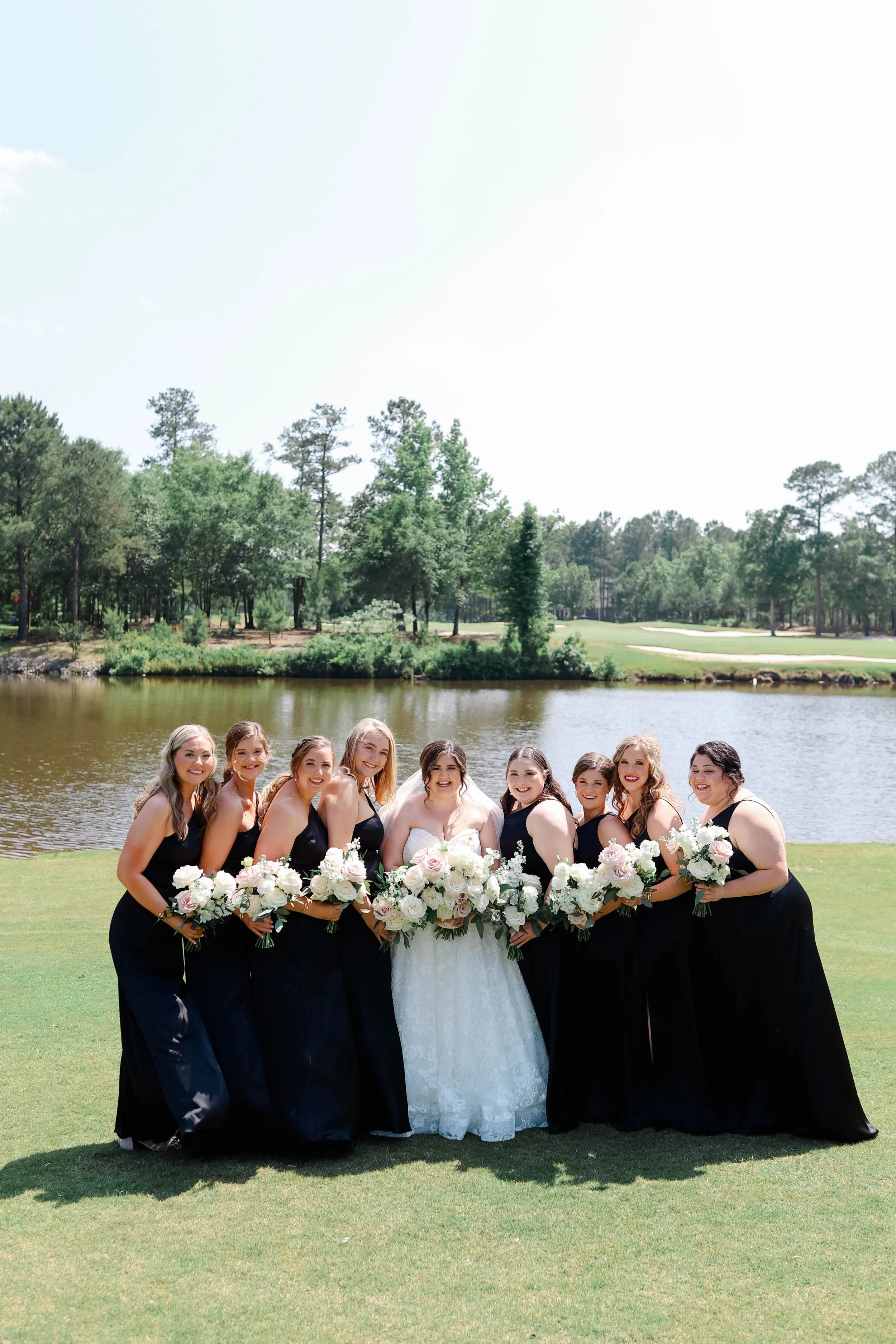 A bride and nine bridesmaids standing outdoors by a pond with trees and grass, holding bouquets of white and blush flowers, dressed in matching black dresses, smiling for a photo on a sunny day.