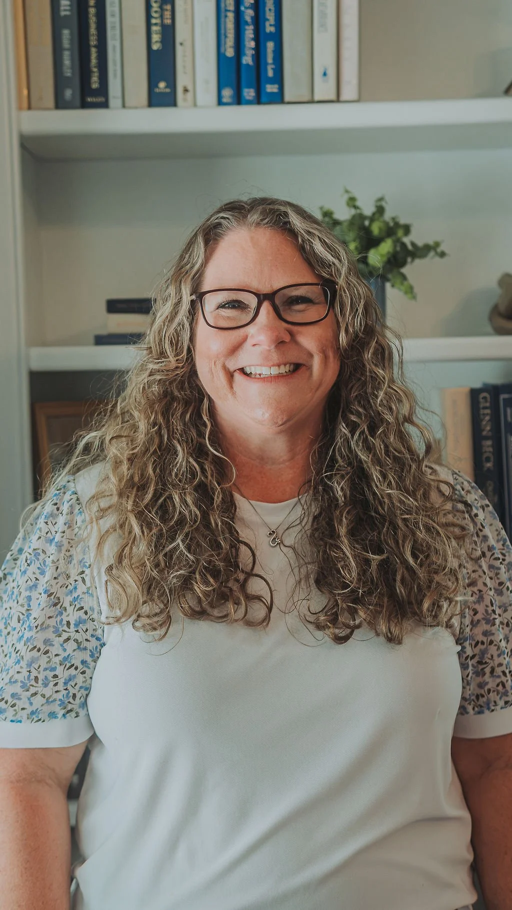 A woman with curly hair and glasses smiling, standing in front of a bookshelf with books and a plant.