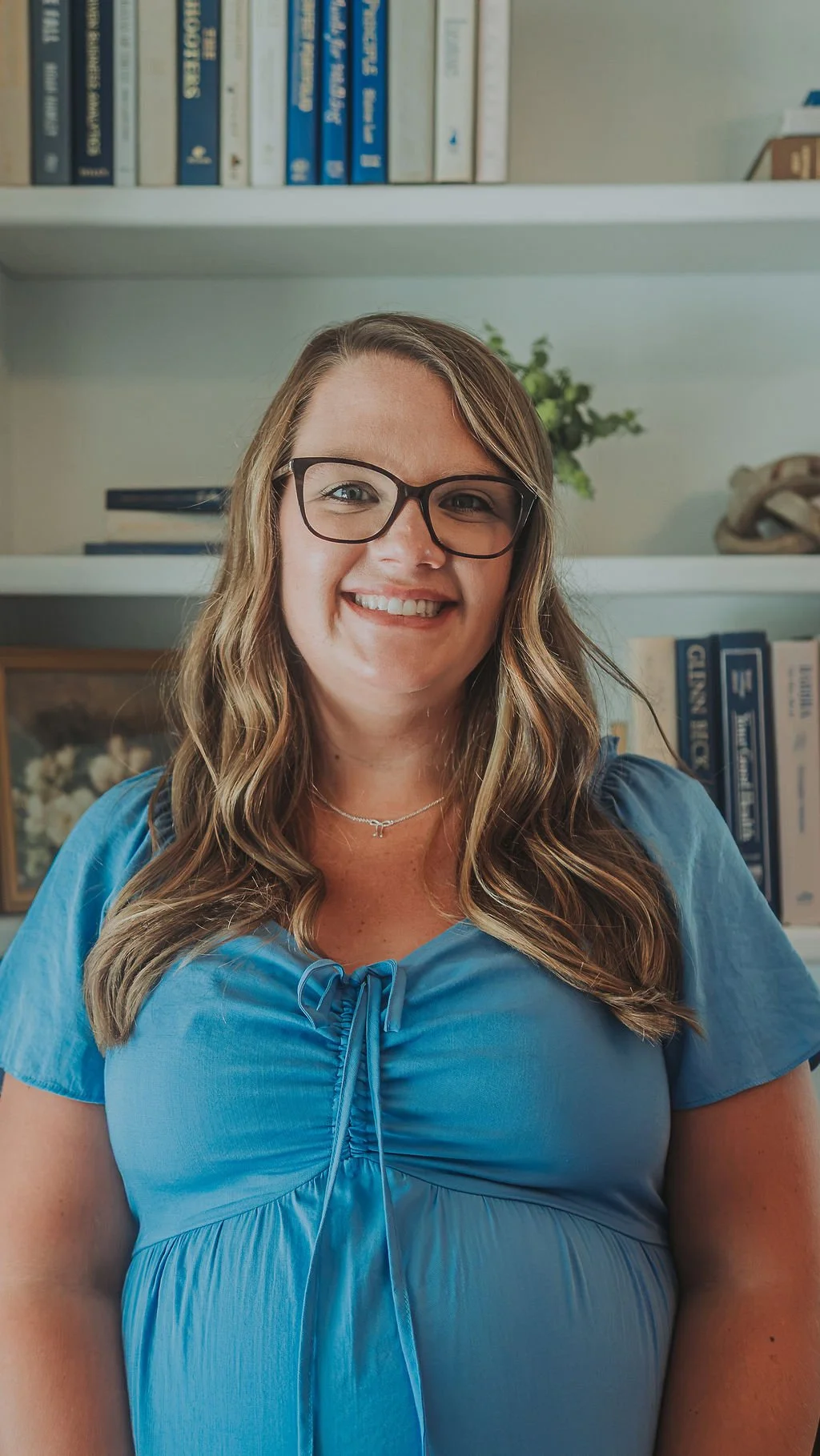 A woman with long wavy hair, glasses, and a blue blouse is smiling in front of a bookshelf with books and decorative items.