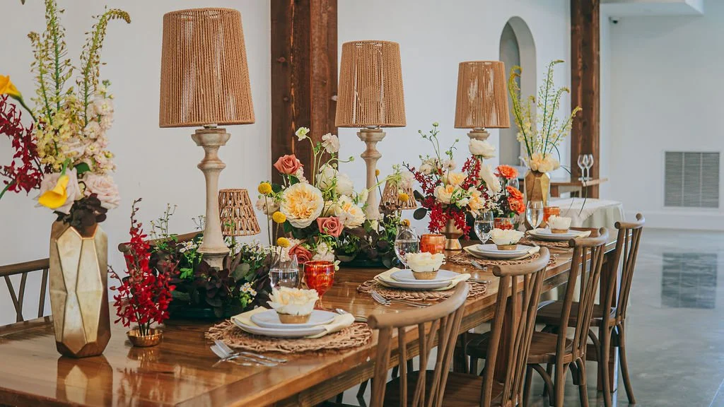 A dining table decorated with floral arrangements, table lamps, and tableware in a well-lit room.