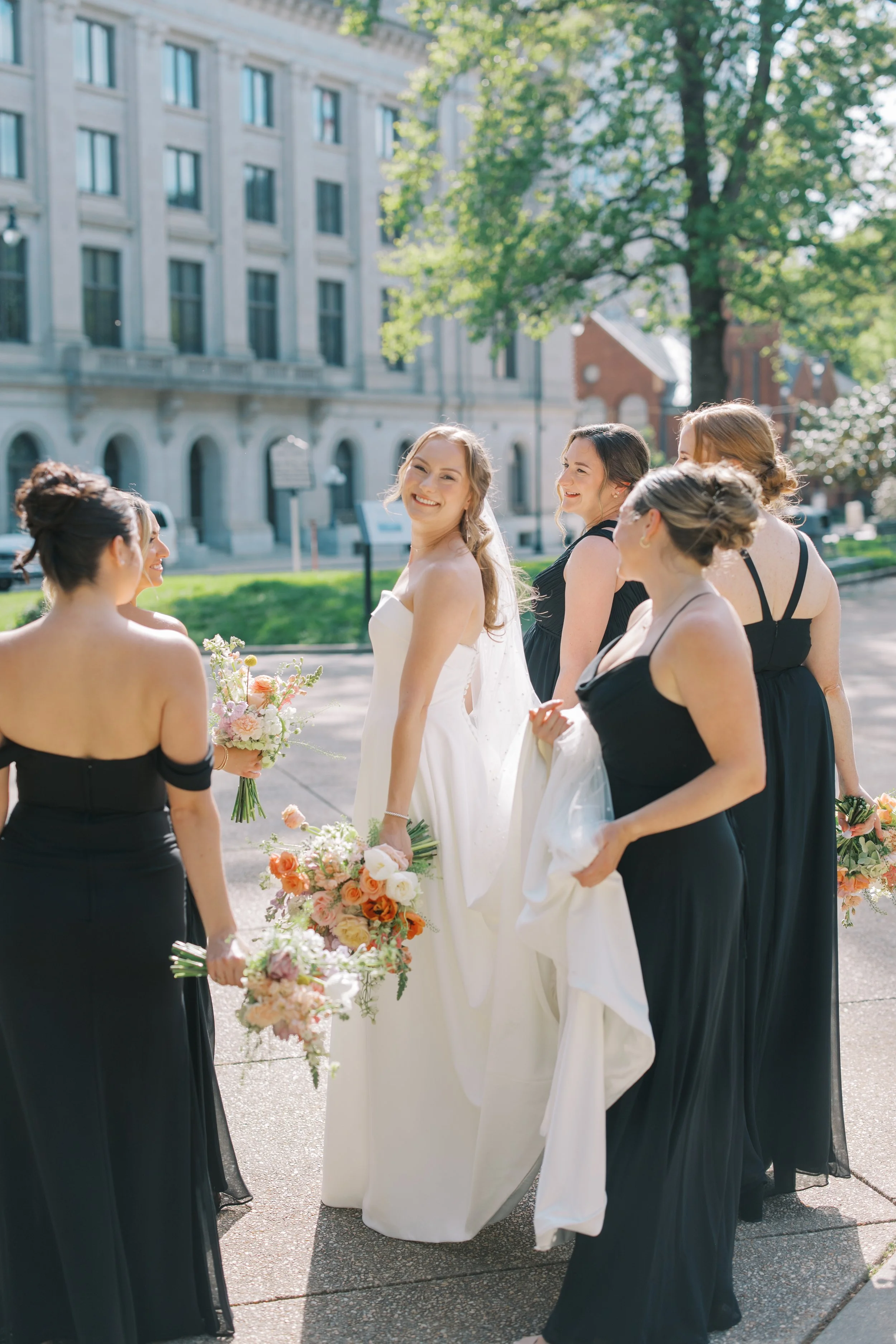 A bride in a white wedding gown smiling at a group of women dressed in black formal dresses outdoors on a sunny day, with a city building and trees in the background.