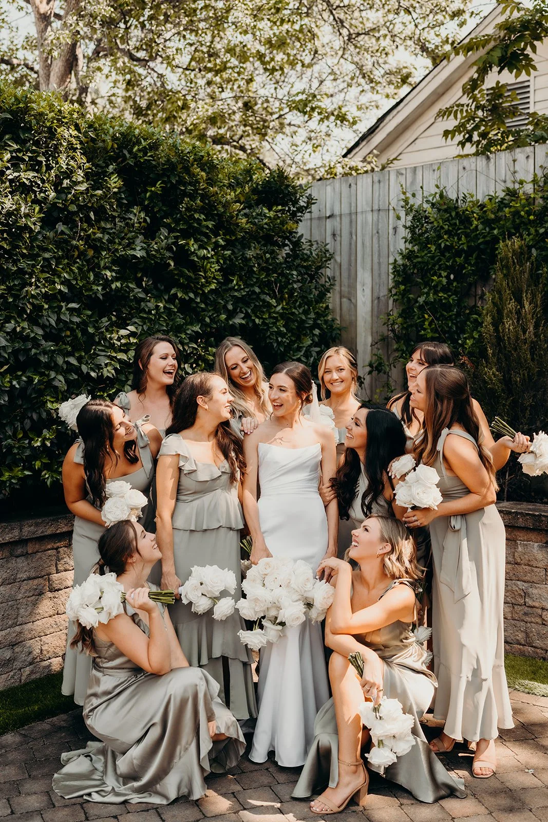 A bride in a white wedding dress surrounded by her bridesmaids in light gray dresses, all holding white bouquets, smiling and laughing in a garden with green bushes and trees.