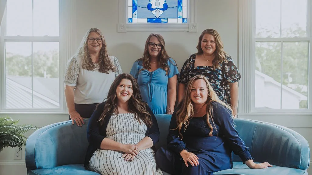 Five women gathered in a well-lit room with large windows, sitting and standing around a blue sofa, smiling for the photo.