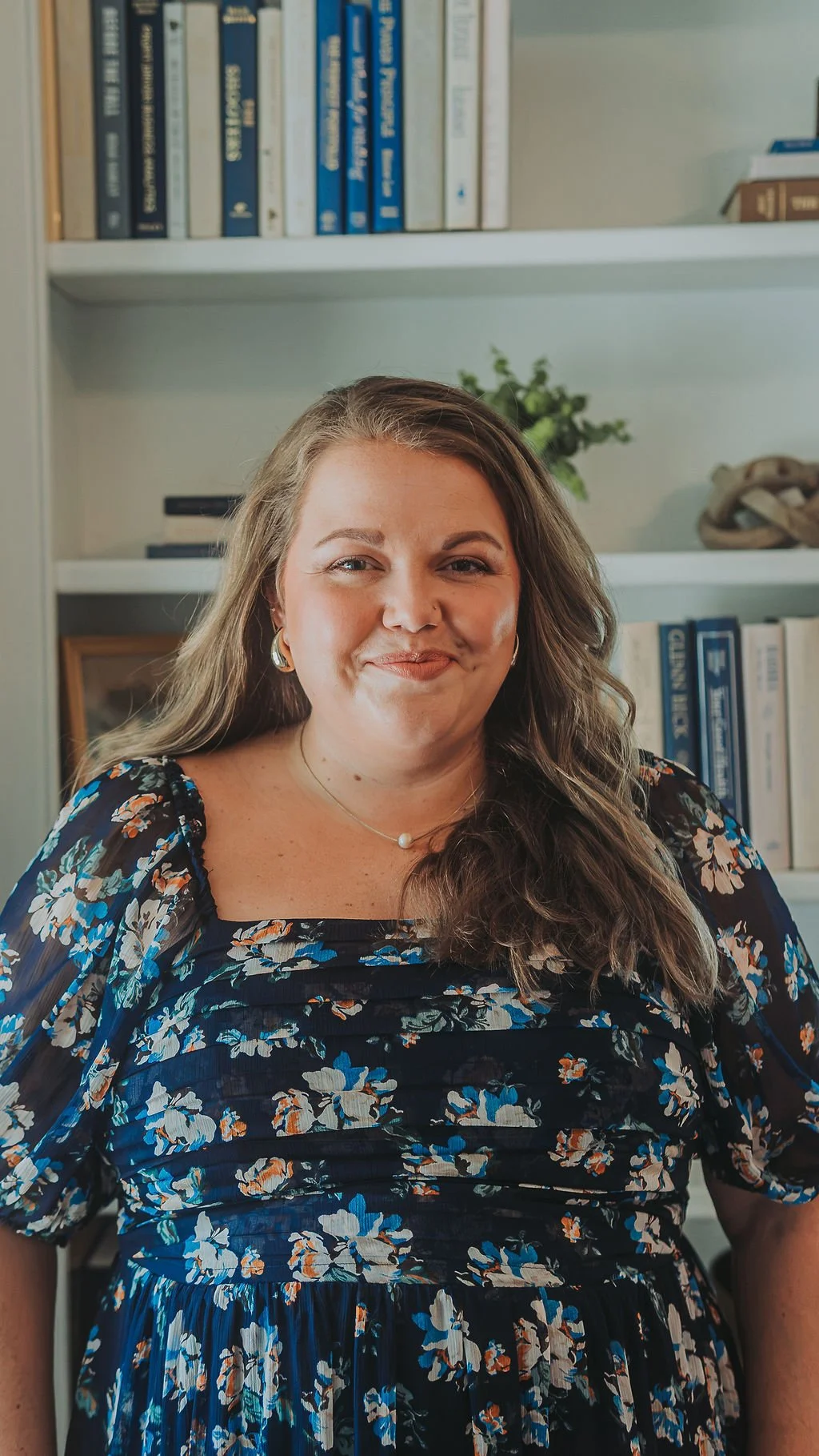 A woman with long wavy brown hair wearing a black floral dress smiles at the camera. She is sitting in front of a bookshelf filled with books and a small potted plant.
