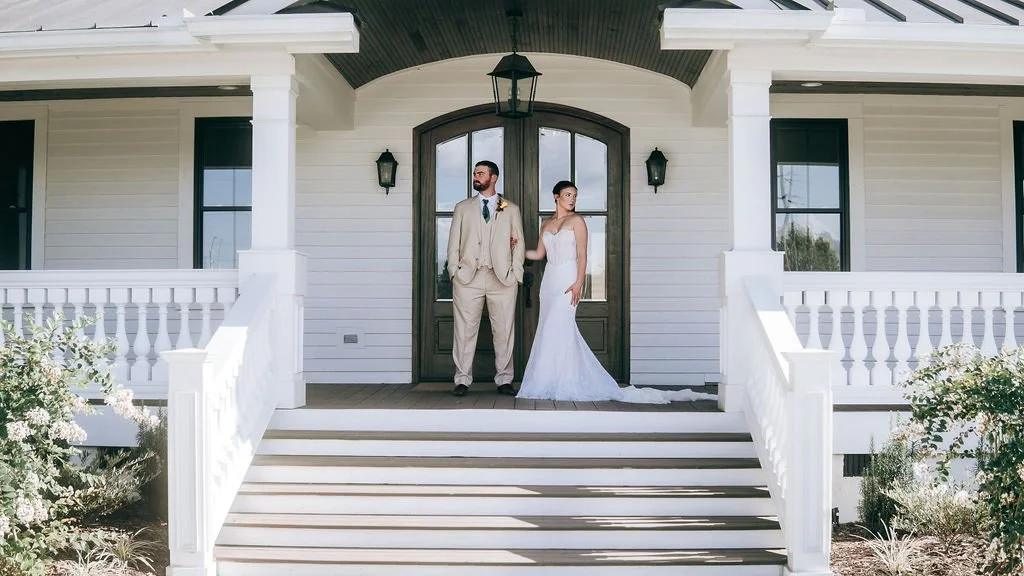 A bride and groom standing on the front porch of a white house with black accents, wedding attire, and floral decorations.