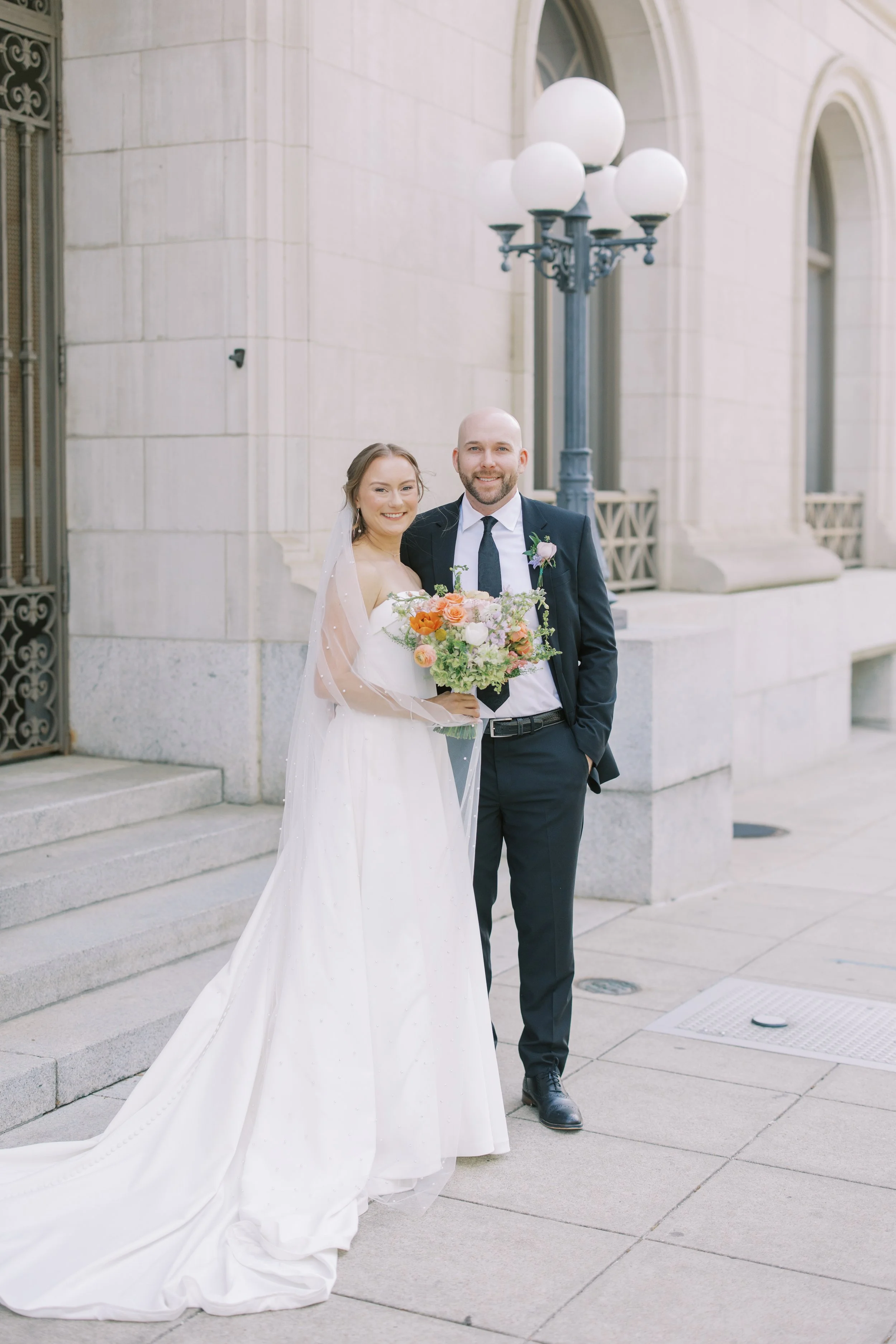 A newly married couple stands outside a building with a stone facade, smiling at the camera. The bride holds a bouquet of colorful flowers and wears a white wedding dress with a veil, while the groom wears a dark suit with a tie and a boutonniere, an