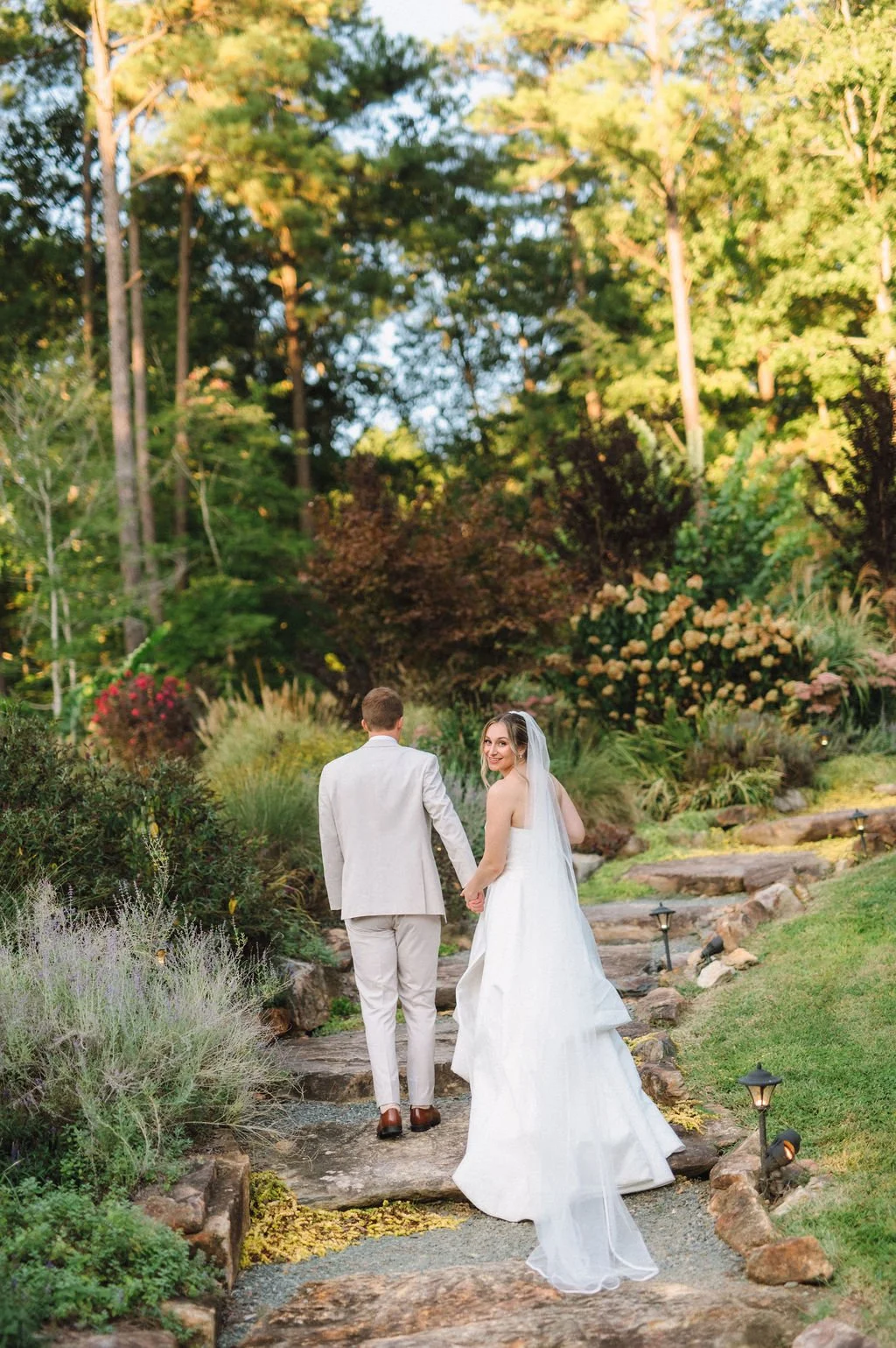 A bride and groom holding hands walking along a stone pathway in a lush garden, with the bride looking back and smiling, surrounded by tall trees and flowering bushes in the late afternoon.
