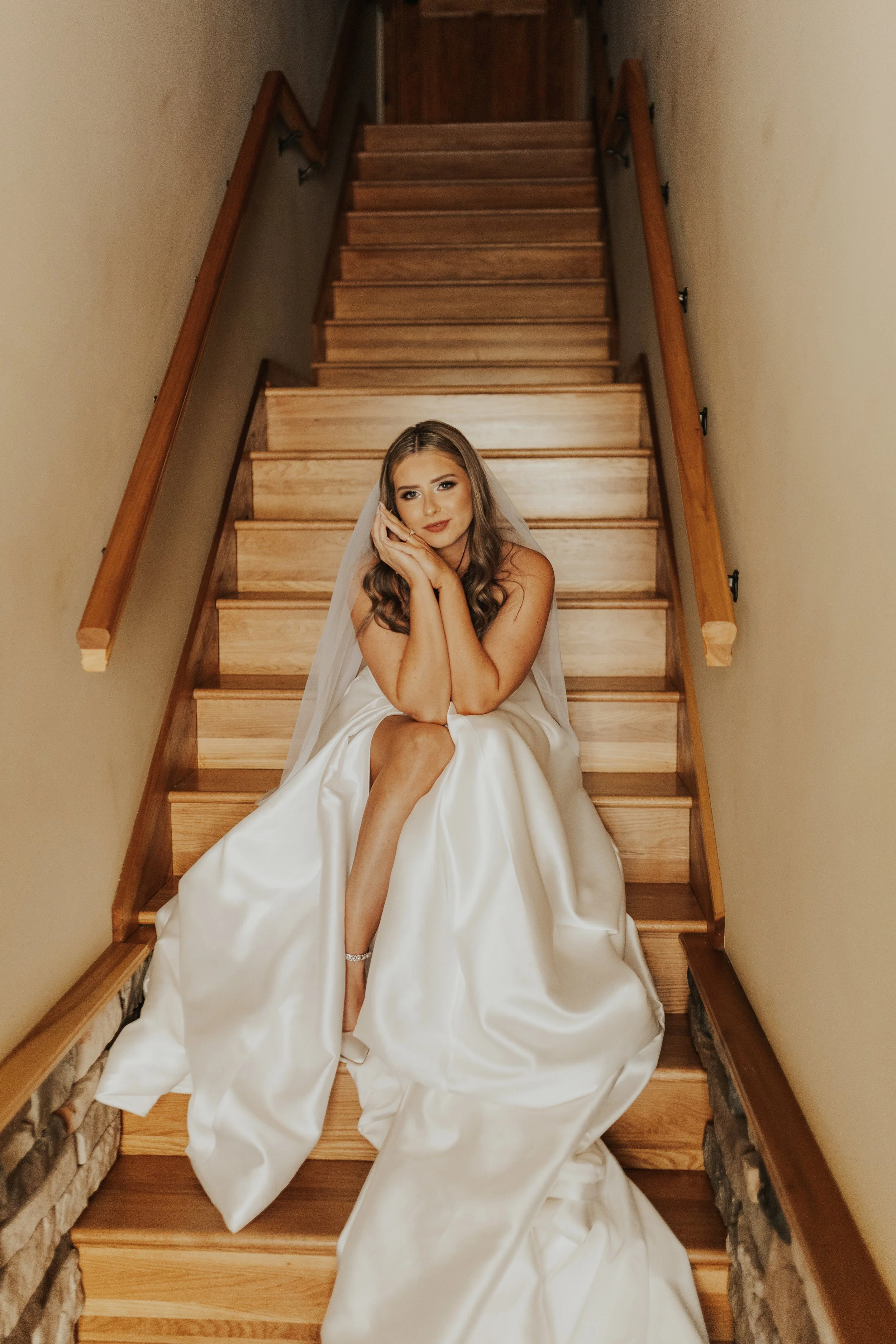 A woman in a wedding dress and veil sitting on wooden stairs, resting her head on her hands, with one leg crossed over the other and the dress spread out on the stairs.