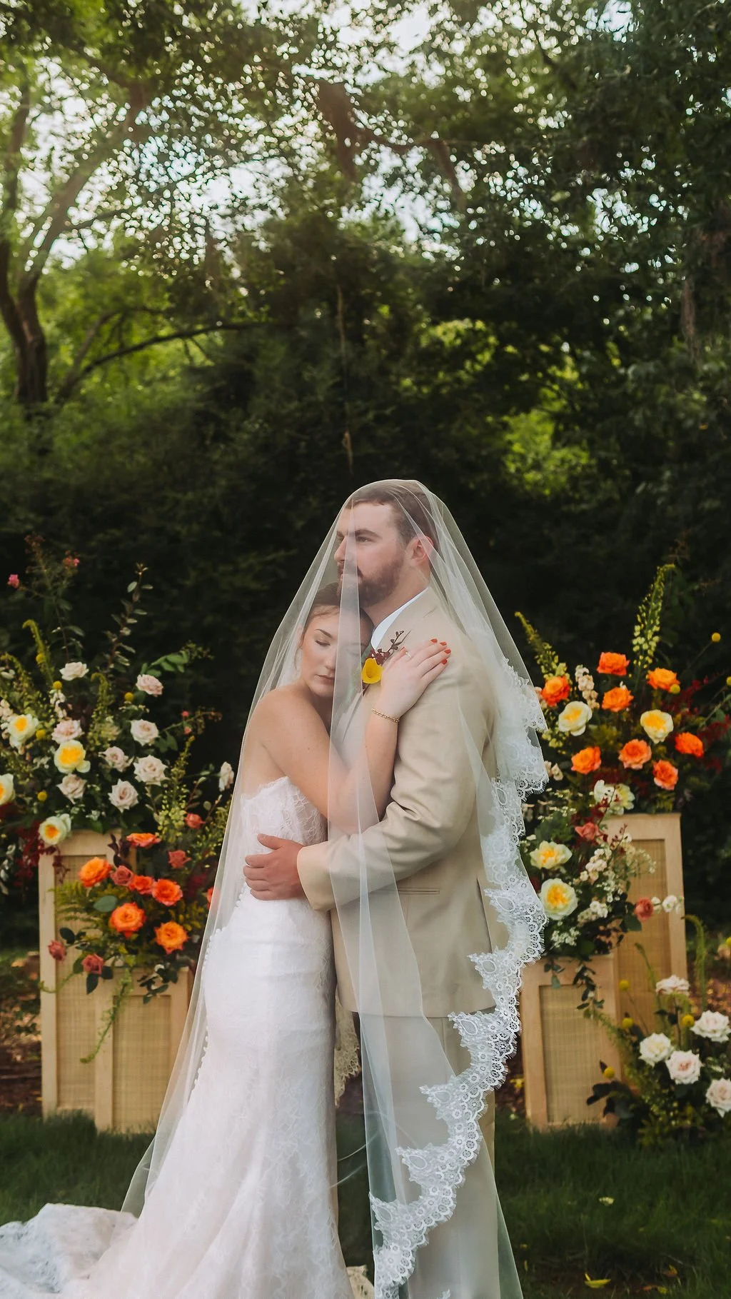 A bride and groom embrace under a veil during an outdoor wedding, with colorful flower arrangements and lush green trees in the background.