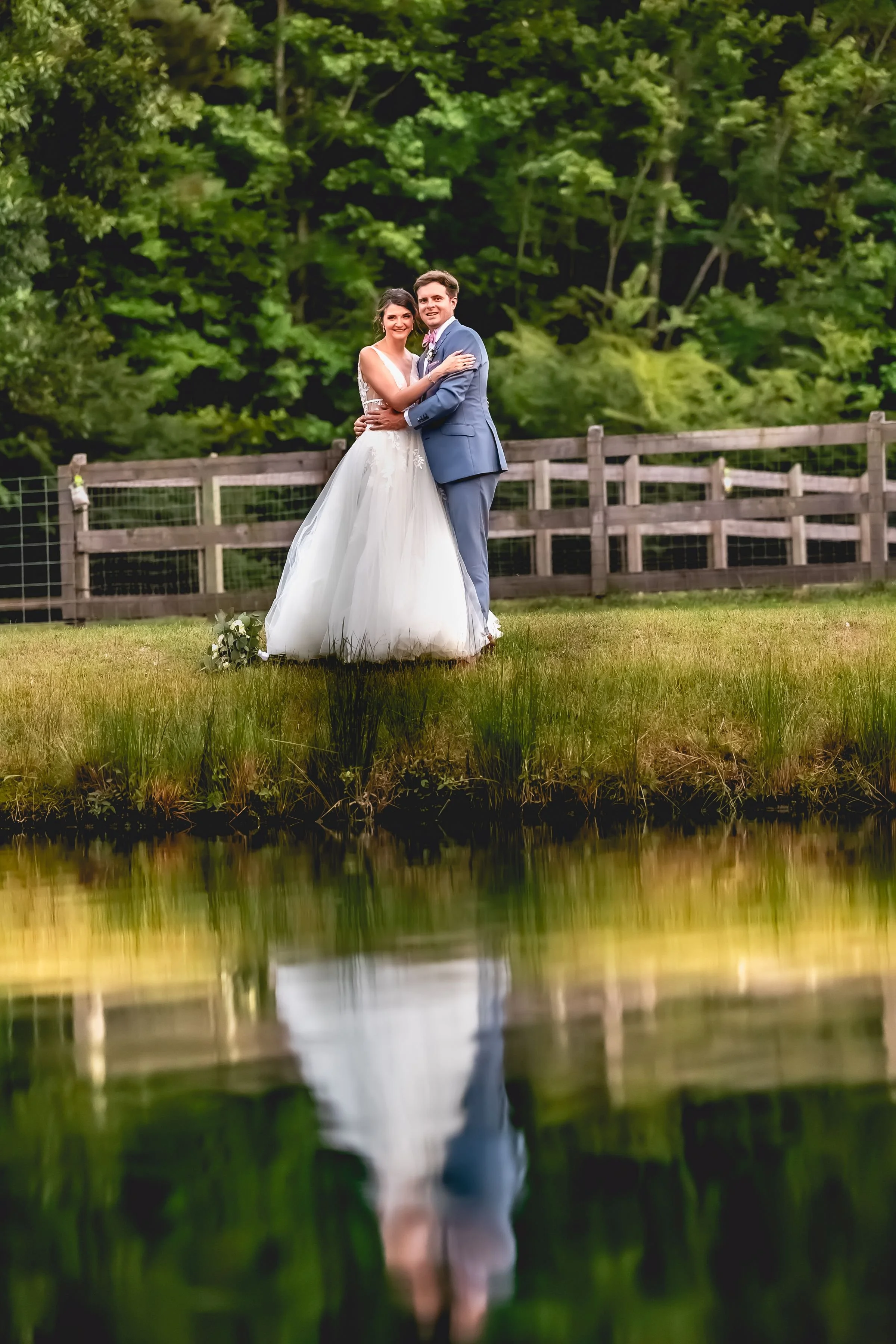 A newlywed couple dressed in wedding attire, standing outdoors in front of a wooden fence, with lush green trees in the background, posing for a wedding photograph.