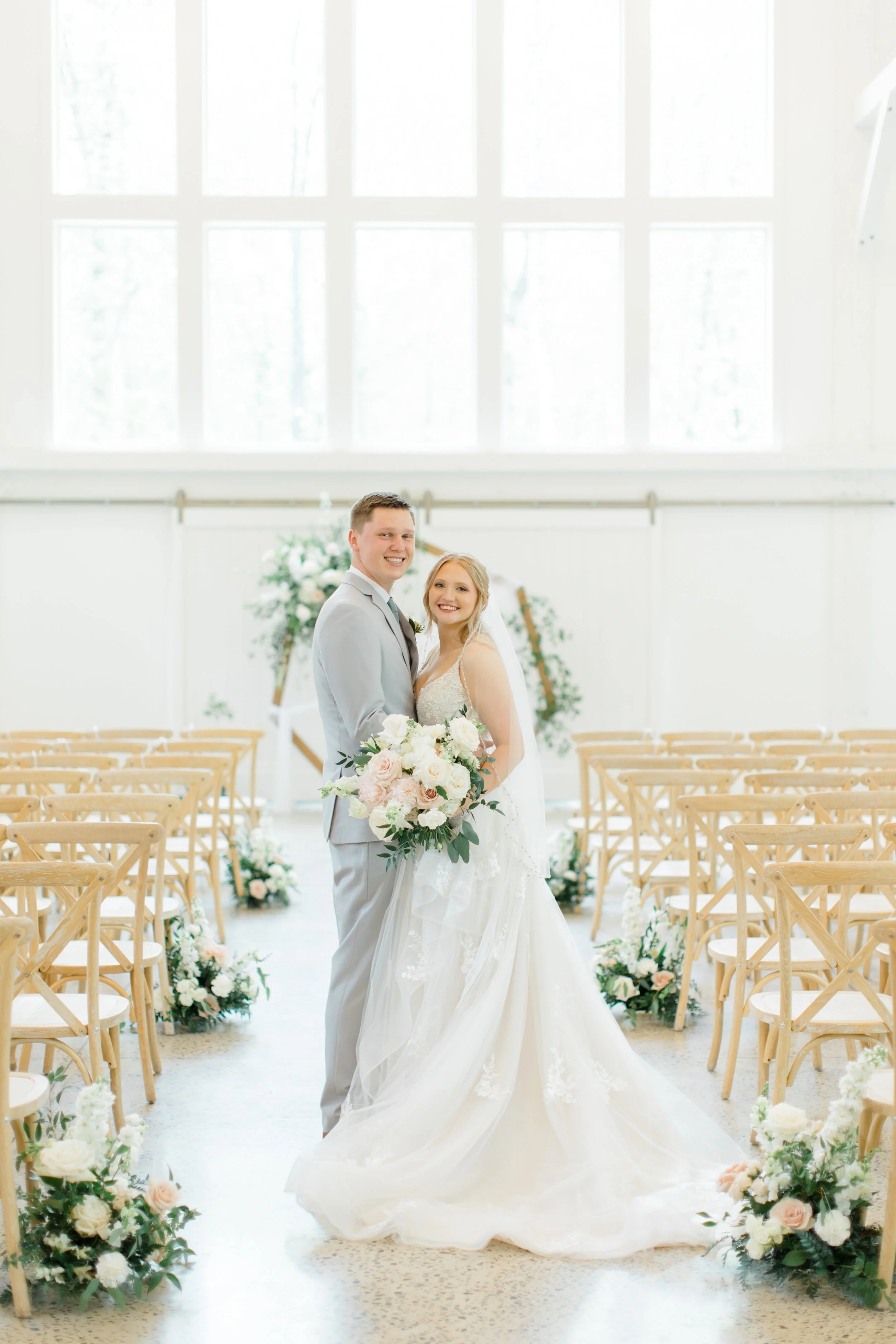 A newlywed couple standing in a bright, decorated wedding venue with empty wooden chairs and floral arrangements, smiling at the camera.