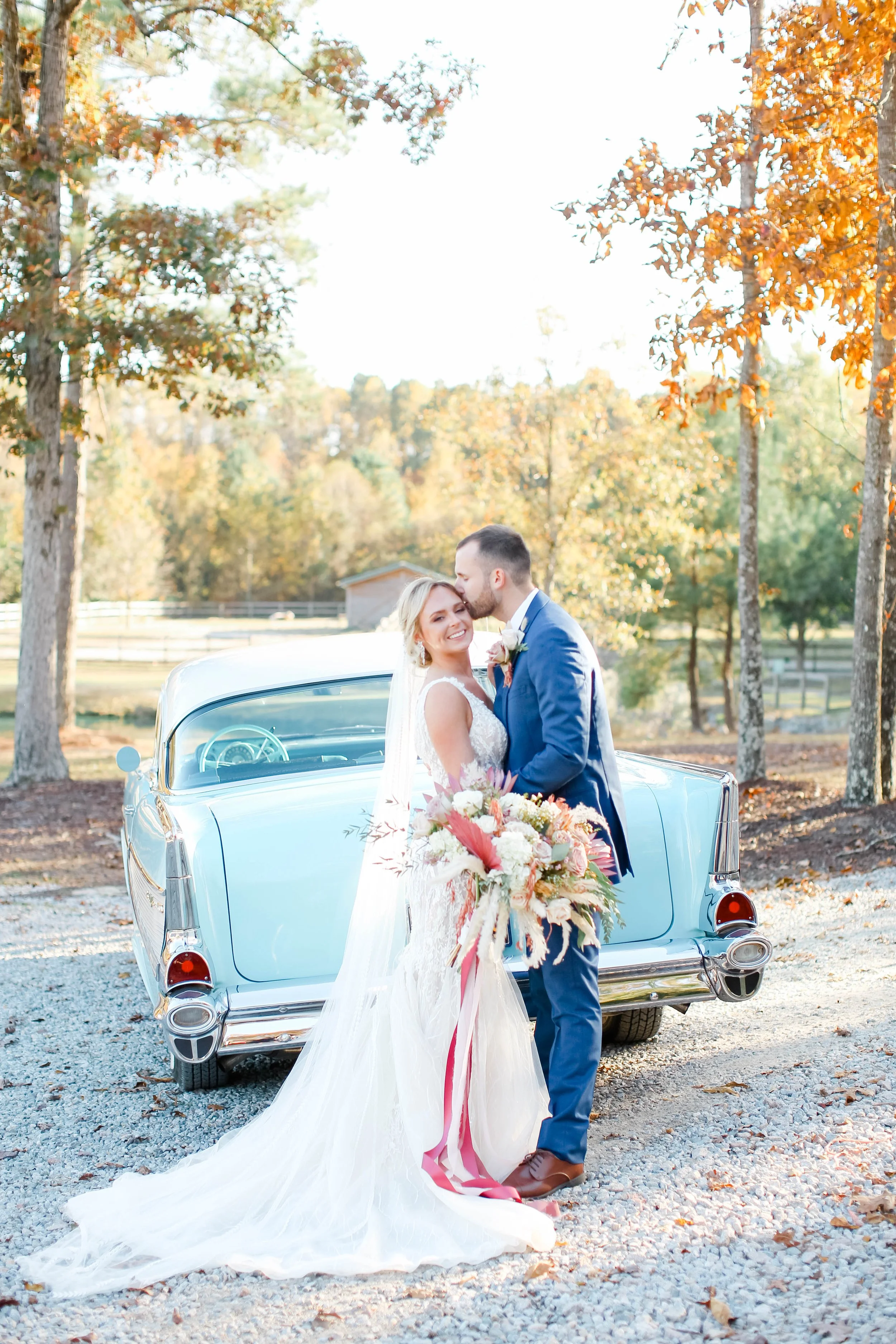 A bride and groom standing in front of a vintage blue car outdoors, surrounded by autumn trees, during a wedding celebration.