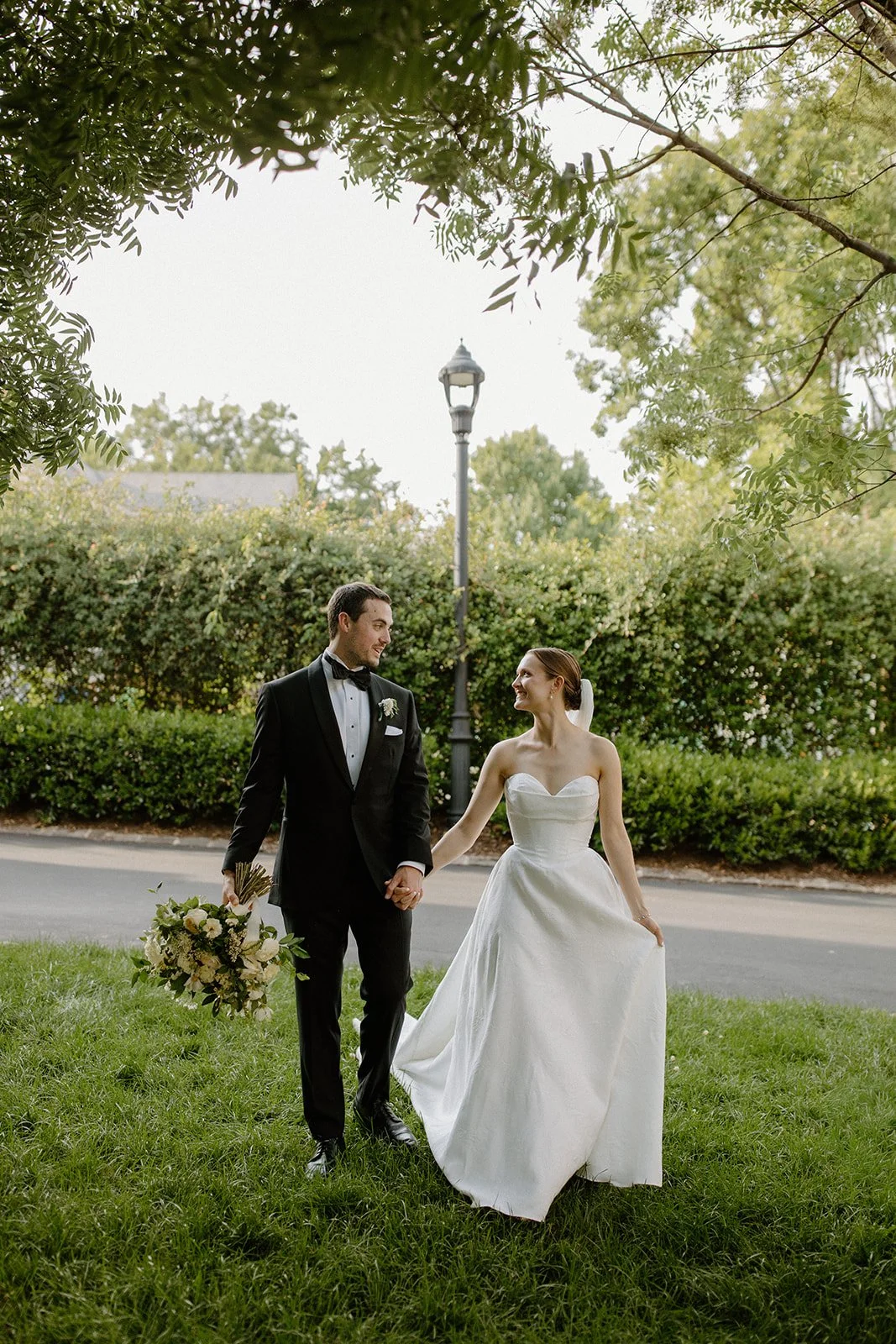 A bride and groom walk hand in hand outdoors on a grassy area, smiling at each other, with trees and a street lamp in the background.
