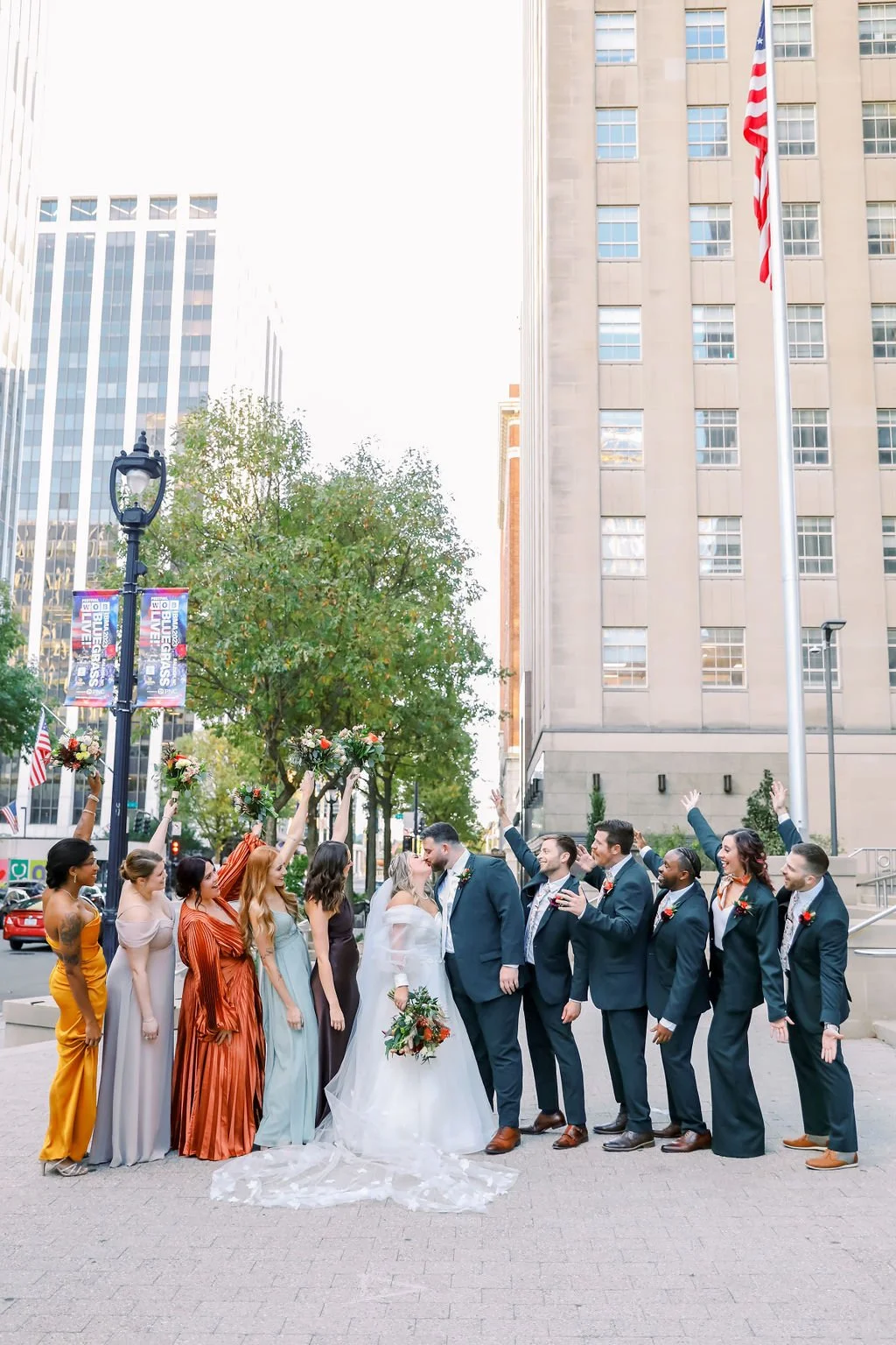A wedding party standing in a city street, with the bride and groom kissing, surrounded by bridesmaids and groomsmen. The bridesmaids hold bouquets with raised arms, and the groomsmen are clapping or raising their hands. Tall buildings and a flagpole