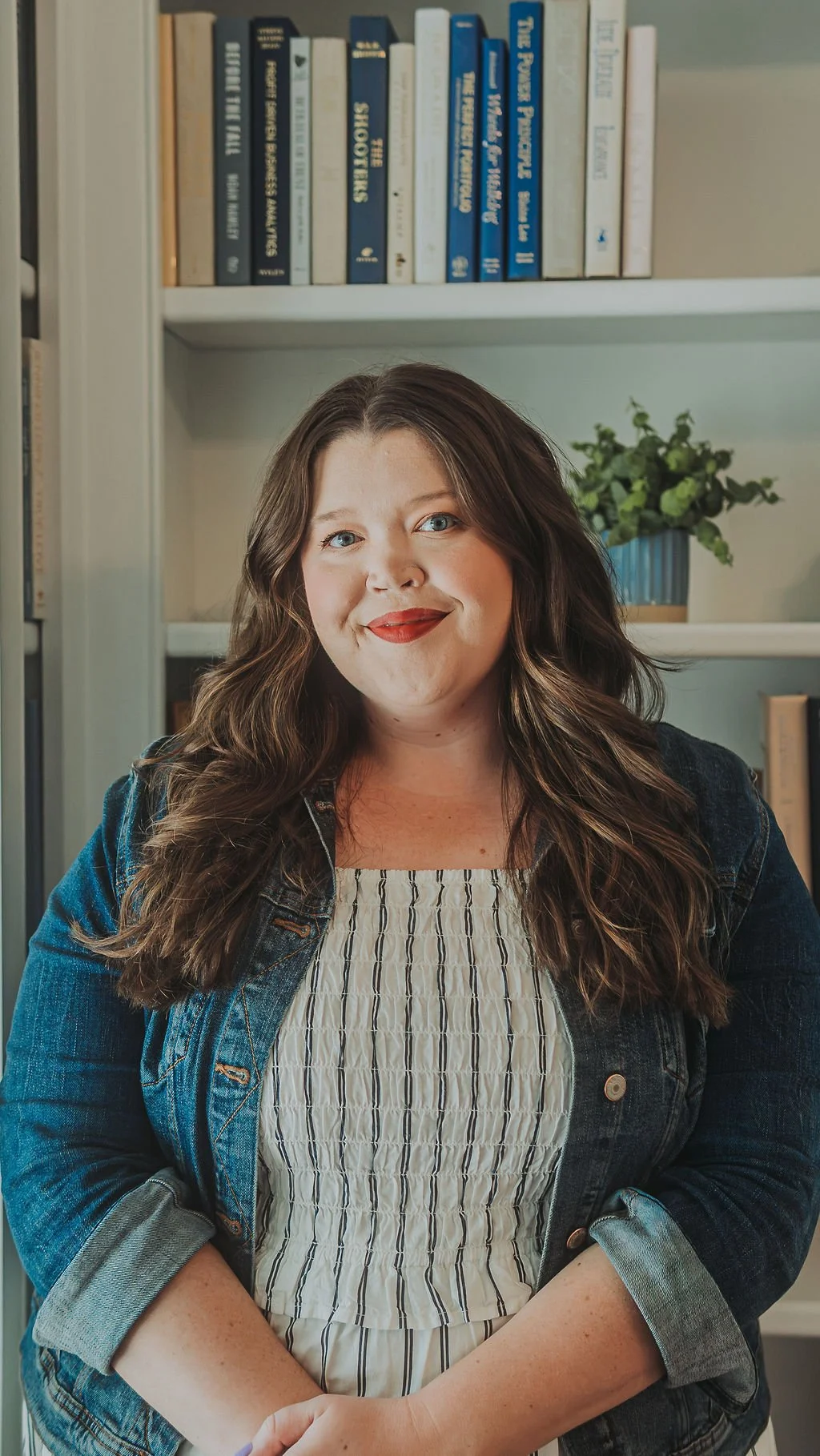 A woman with long wavy brown hair and blue eyes smiling in front of a bookshelf with books and a potted plant.