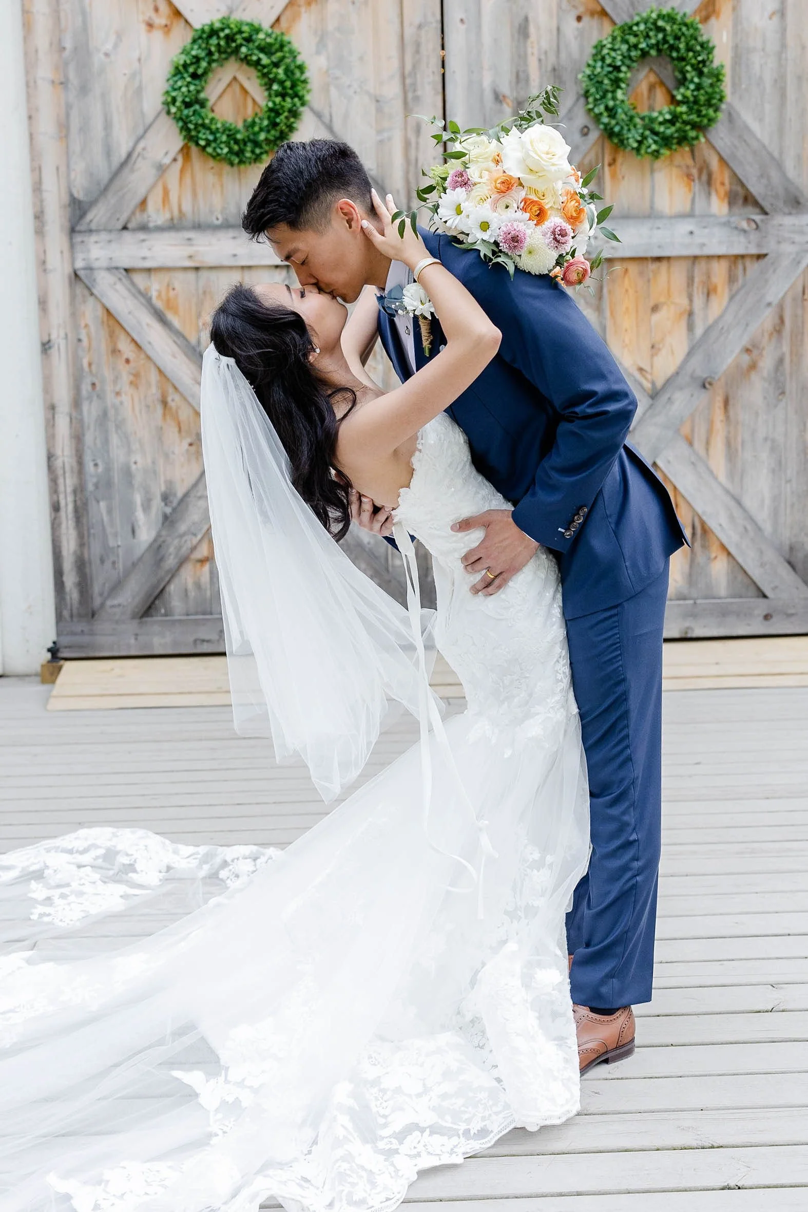 A wedding couple sharing a kiss outdoors, with the groom in a navy blue suit and the bride in a white wedding dress with a long veil, standing in front of wooden doors decorated with green wreaths.