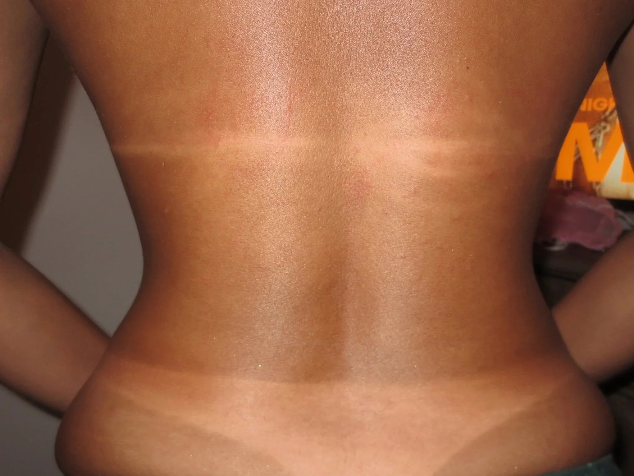 Close-up of a person's shirtless upper torso and shoulders, with tan skin and a background featuring orange and black packaging.