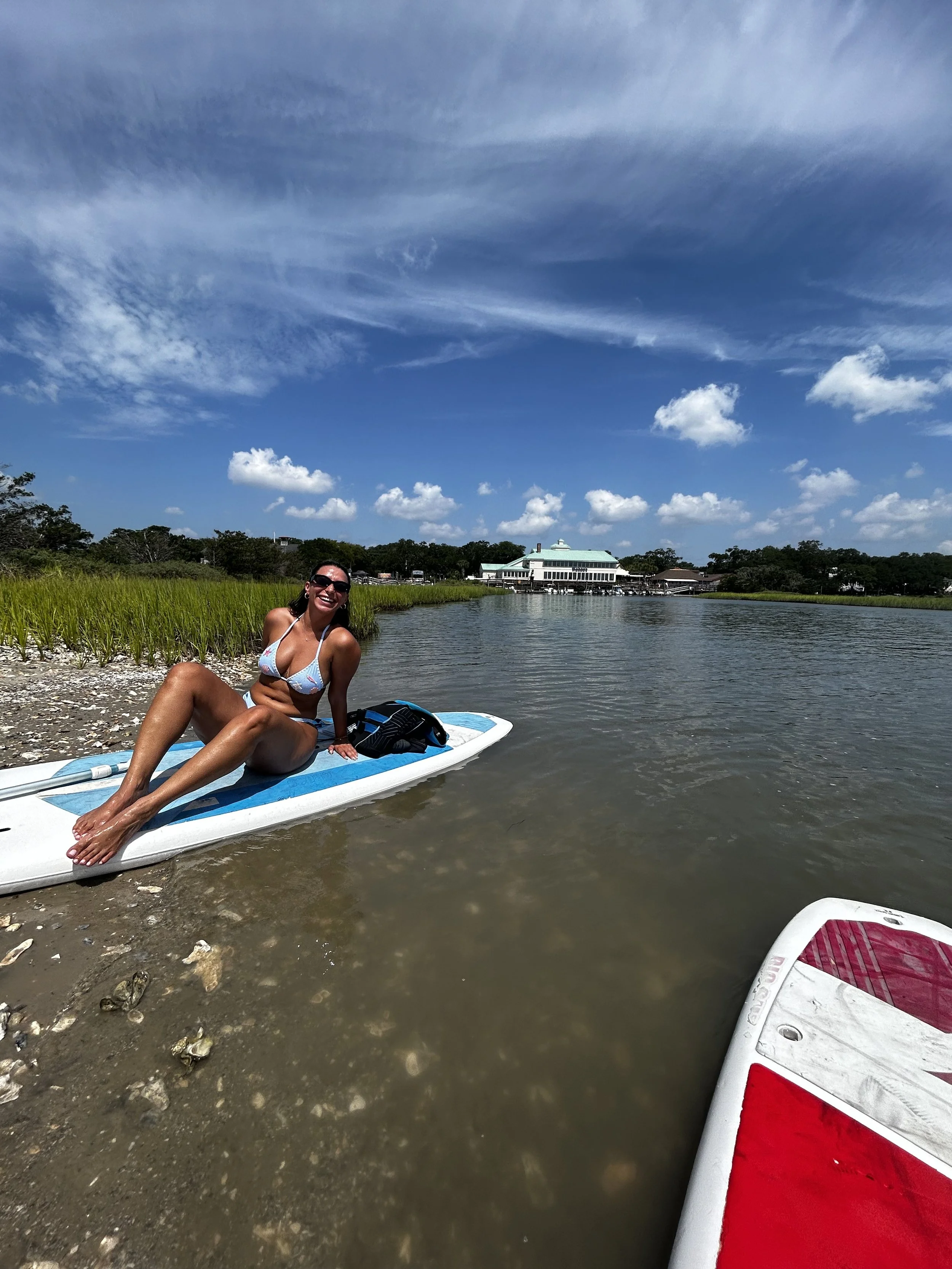 A woman in a bikini sitting on a paddleboard near a sandy and rocky shoreline with a building and trees in the background, under a partly cloudy sky.