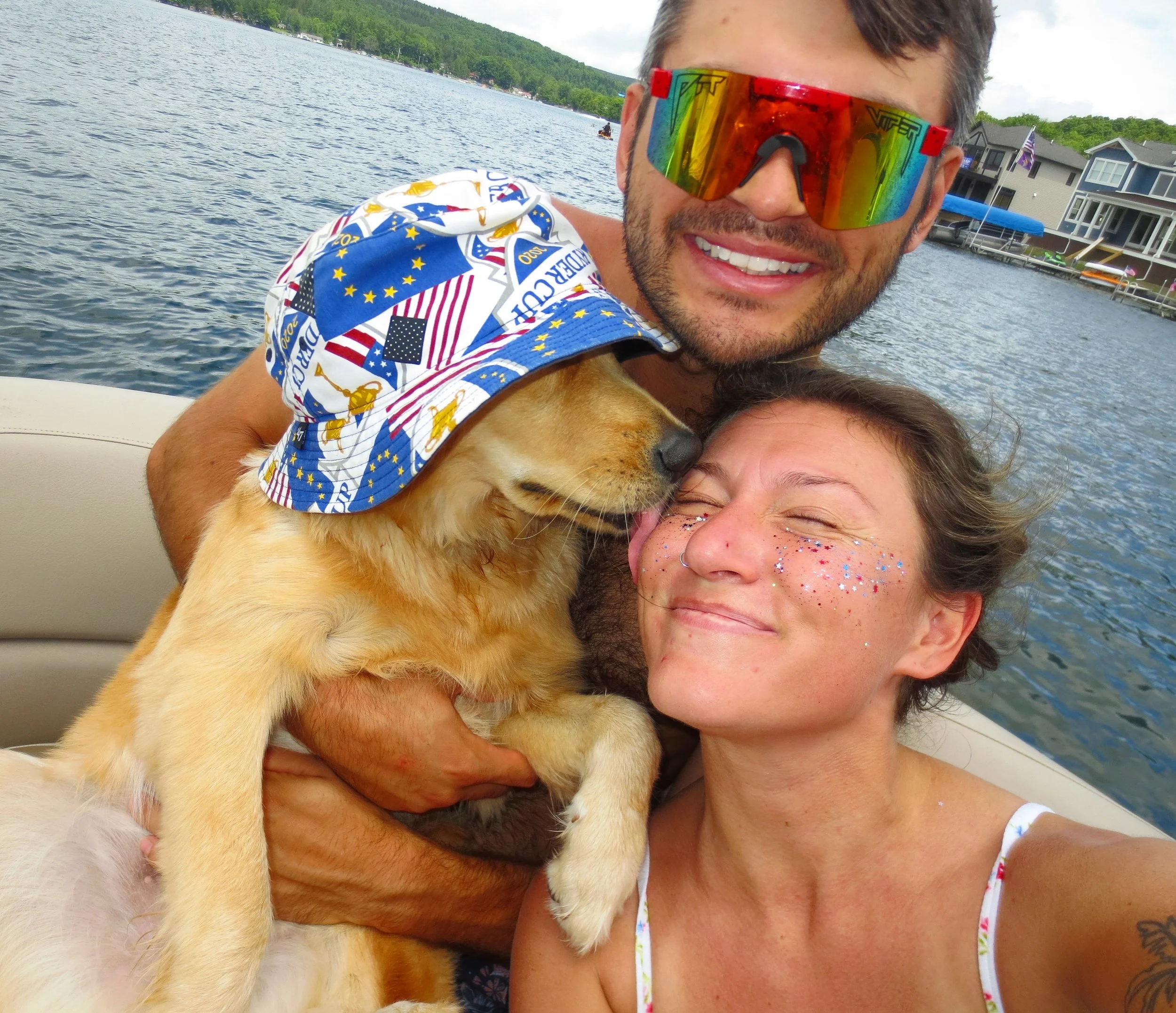 A man, woman, and dog taking a selfie on a boat by a lake. The man is wearing large rainbow-colored sunglasses and a shirt with a space-themed pattern. The woman has colorful glitter on her face, and the dog is wearing a bandana. Houses and trees are visible in the background along the shoreline.