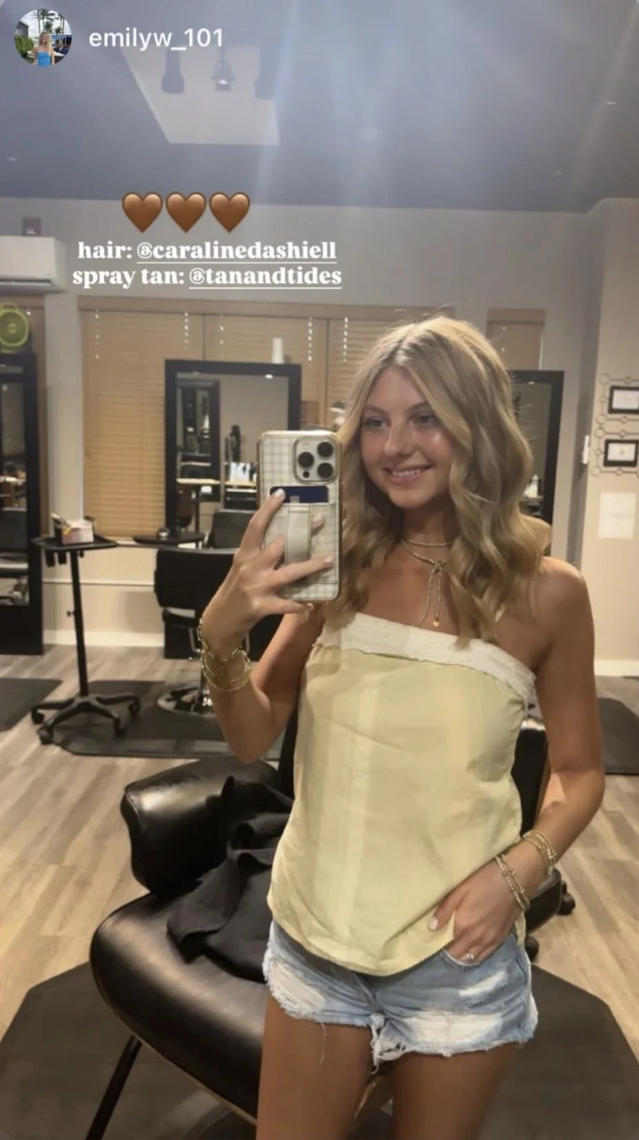 A young woman with wavy blonde hair taking a selfie in a hair salon, smiling, wearing a yellow strapless top and distressed denim shorts, jewelry on her wrists and neck, with salon chairs, mirrors, and equipment in the background.