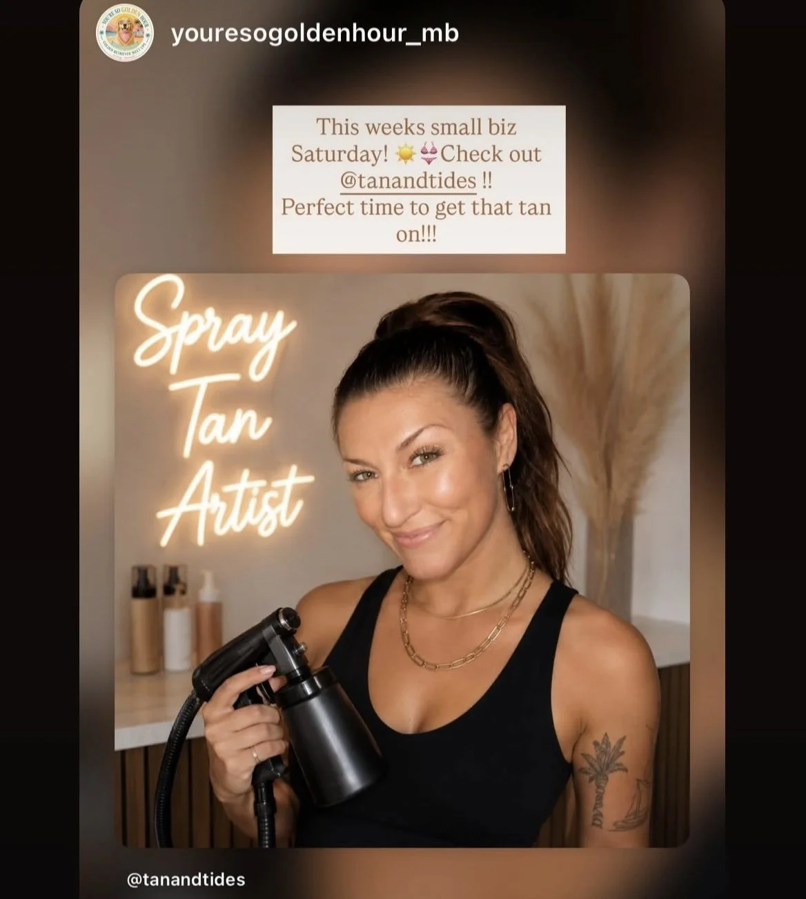 A woman with dark hair tied in a ponytail is holding a spray tanning machine and smiling at the camera. Behind her is a warm-toned background with decor, and a neon sign that says 'Spray Tan Artist'. There is also a text overlay promoting a tanning service for Saturday.