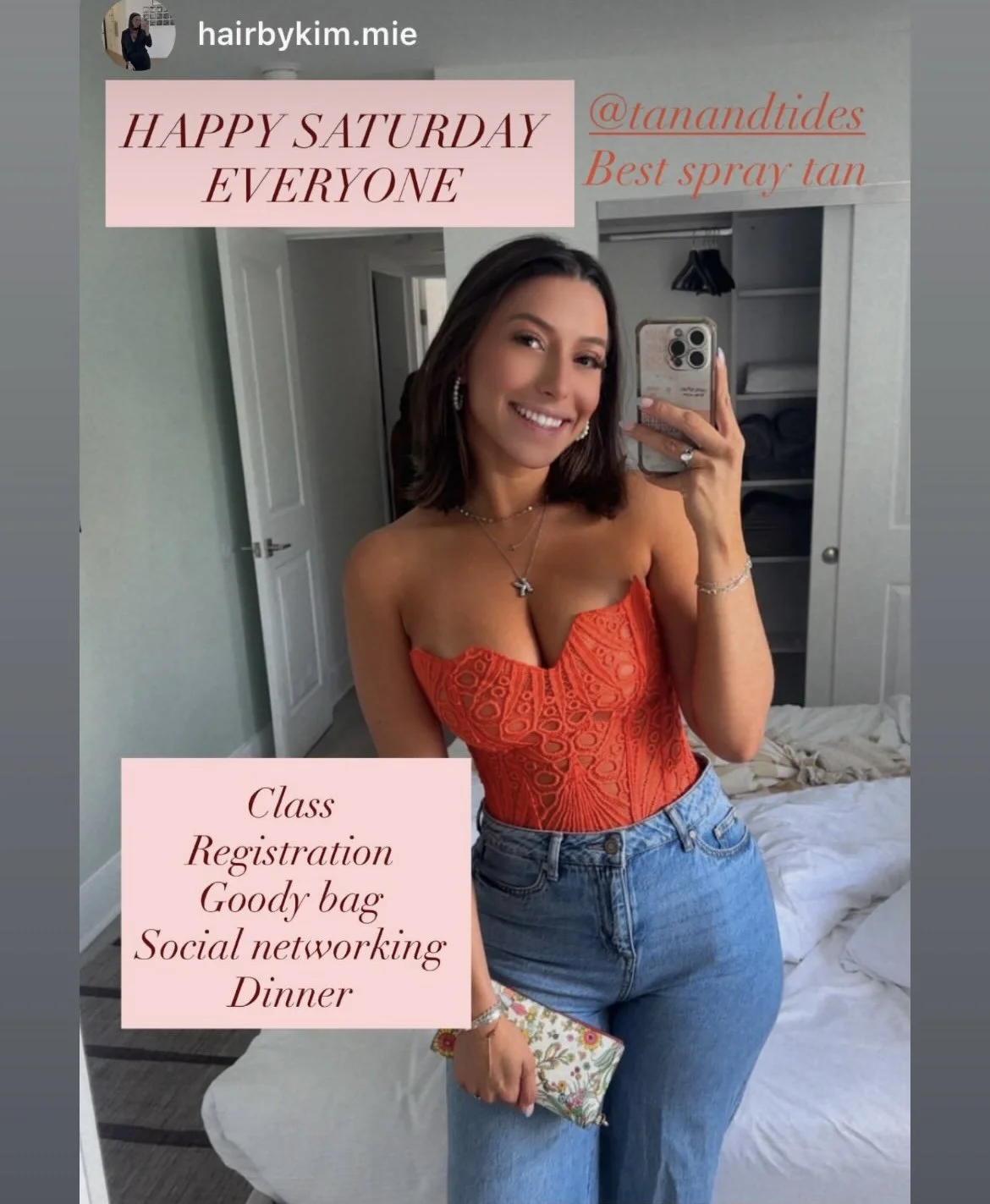 A woman taking a mirror selfie, smiling, wearing an orange strapless top and blue jeans, in a bedroom with an open closet and a bed with white sheets.