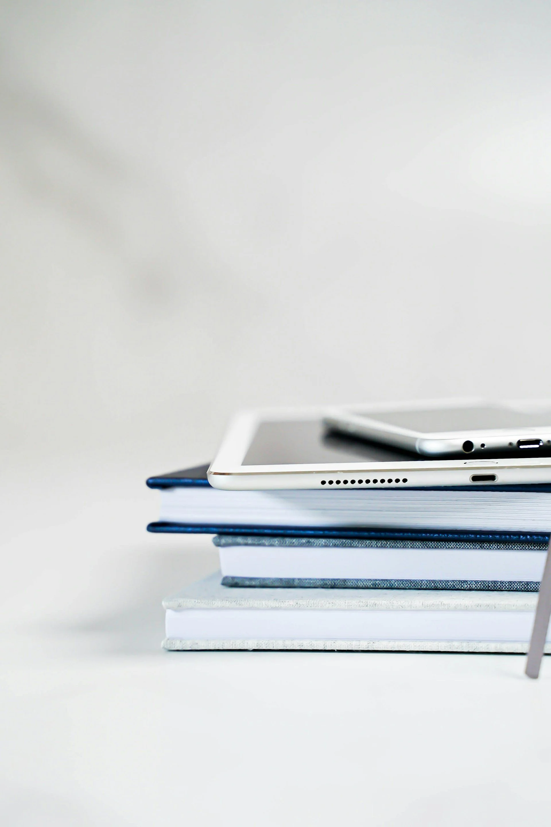 Stack of books with a notebook, a tablet, and a smartphone on top against a plain background.
