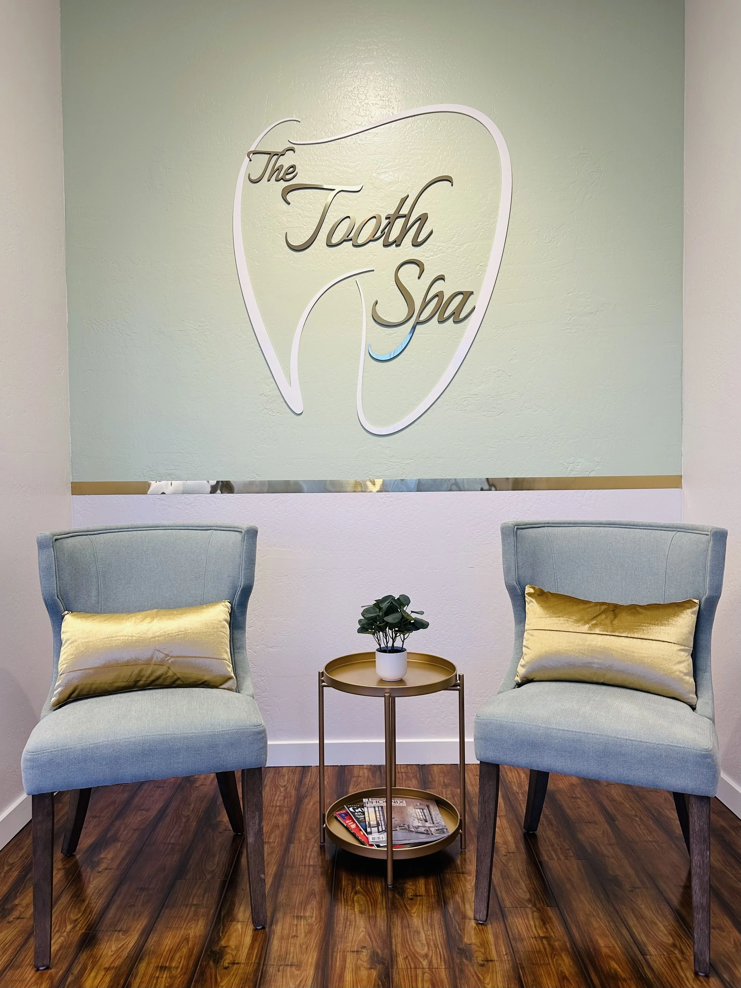A waiting area with two chairs, accent gold pillows, round side table with a potted plant, and a wall sign that reads "The Tooth Spa."