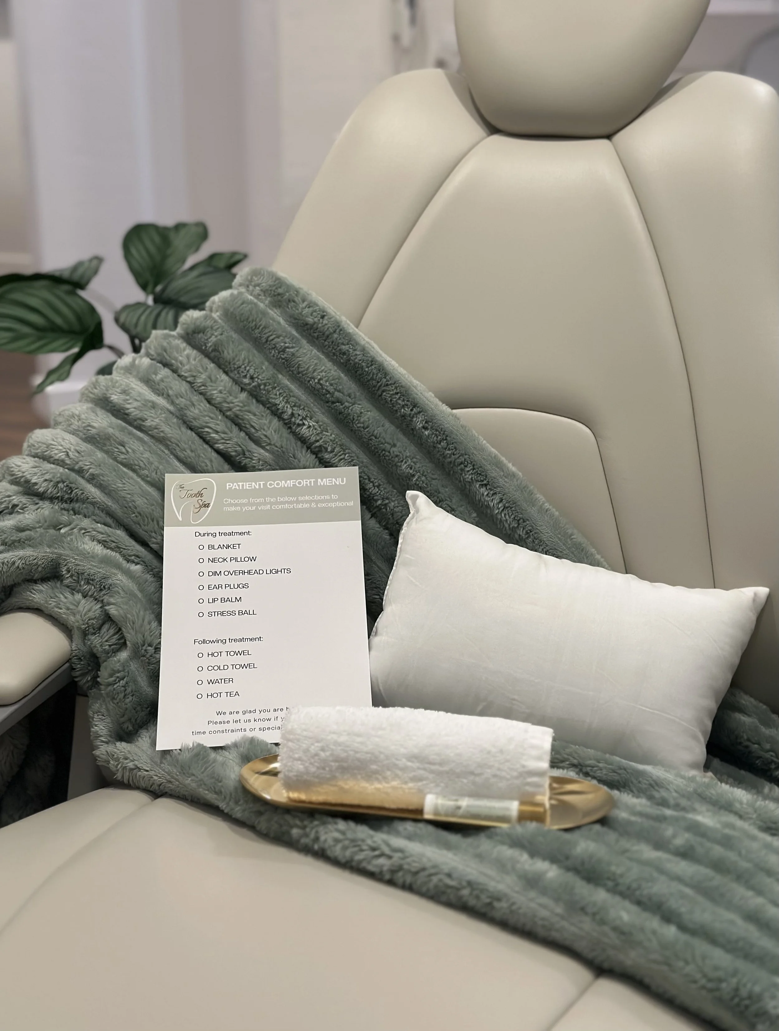 A plush blanket and pillow are on a spa dental chair with a patient comfort menu next to them. The background shows part of a white wall and some greenery.