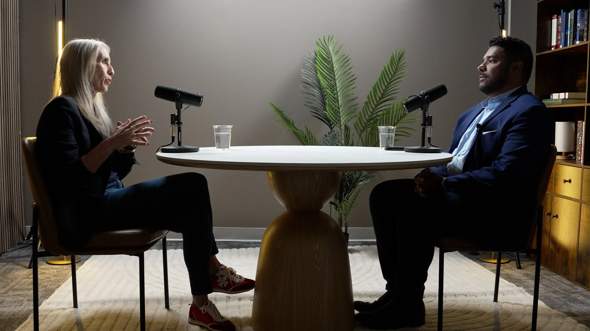 A woman and a man seated across from each other at a round table with microphones, glasses of water, and a plant in the background, engaged in a conversation or an interview.