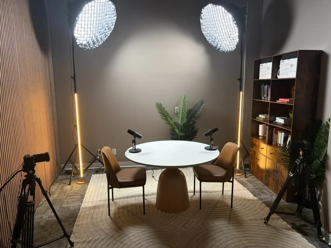 A professional podcast or interview setup with a round white table, two brown chairs, two microphones, and professional lighting in a cozy room with wood paneling and a bookshelf.
