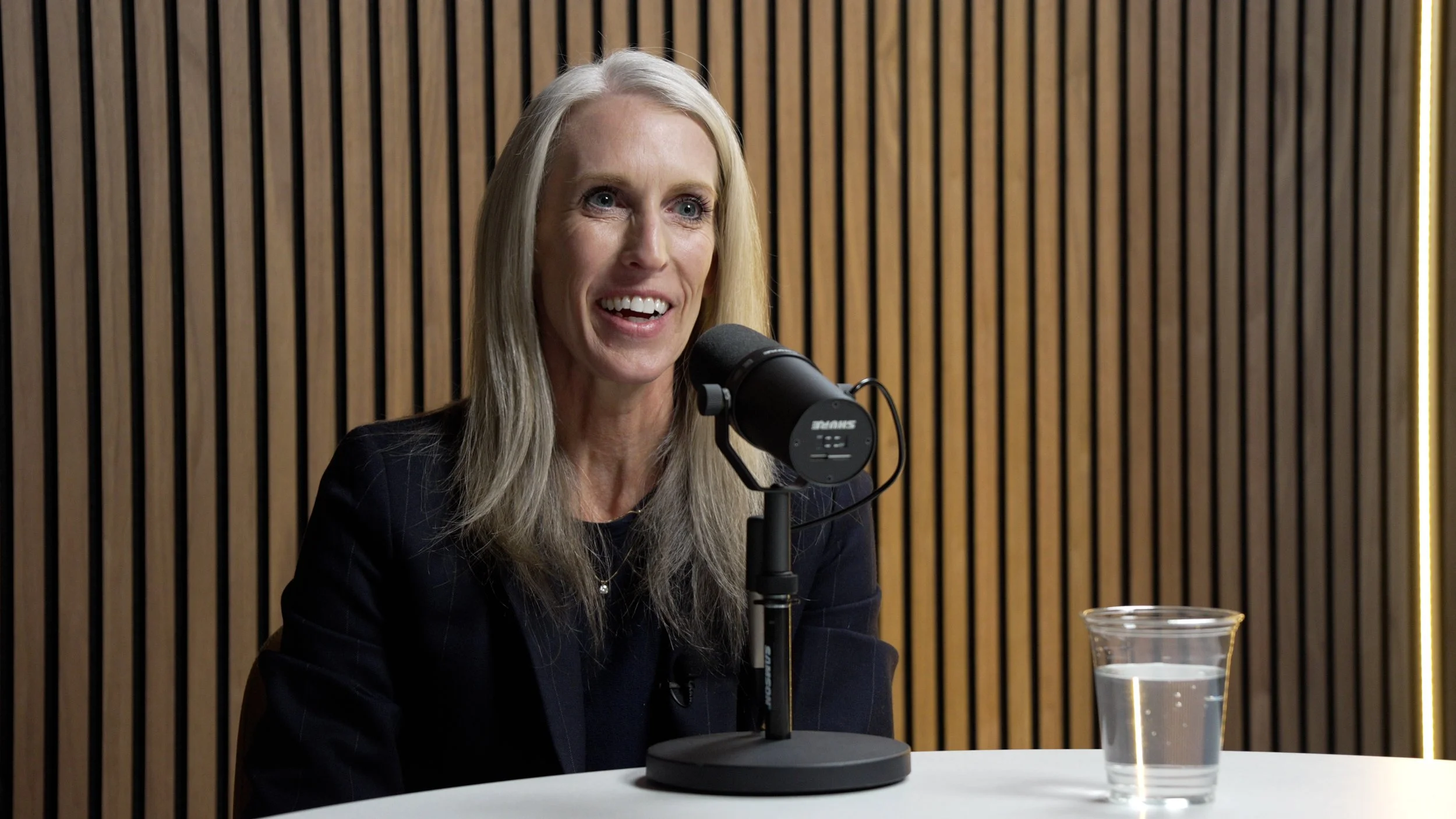 A woman with long blonde hair smiling and speaking into a microphone at a panel discussion or podcast studio with wooden slats background and a glass of water on the table.