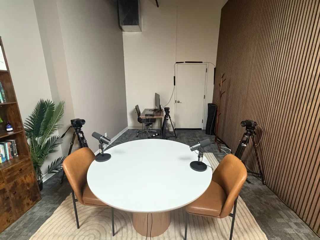 A modern podcast recording studio with a round white table, two brown leather chairs, three microphones, and two cameras on tripods. There is a plant and a wooden bookshelf with books, with a wooden wall on one side and a white wall on another.