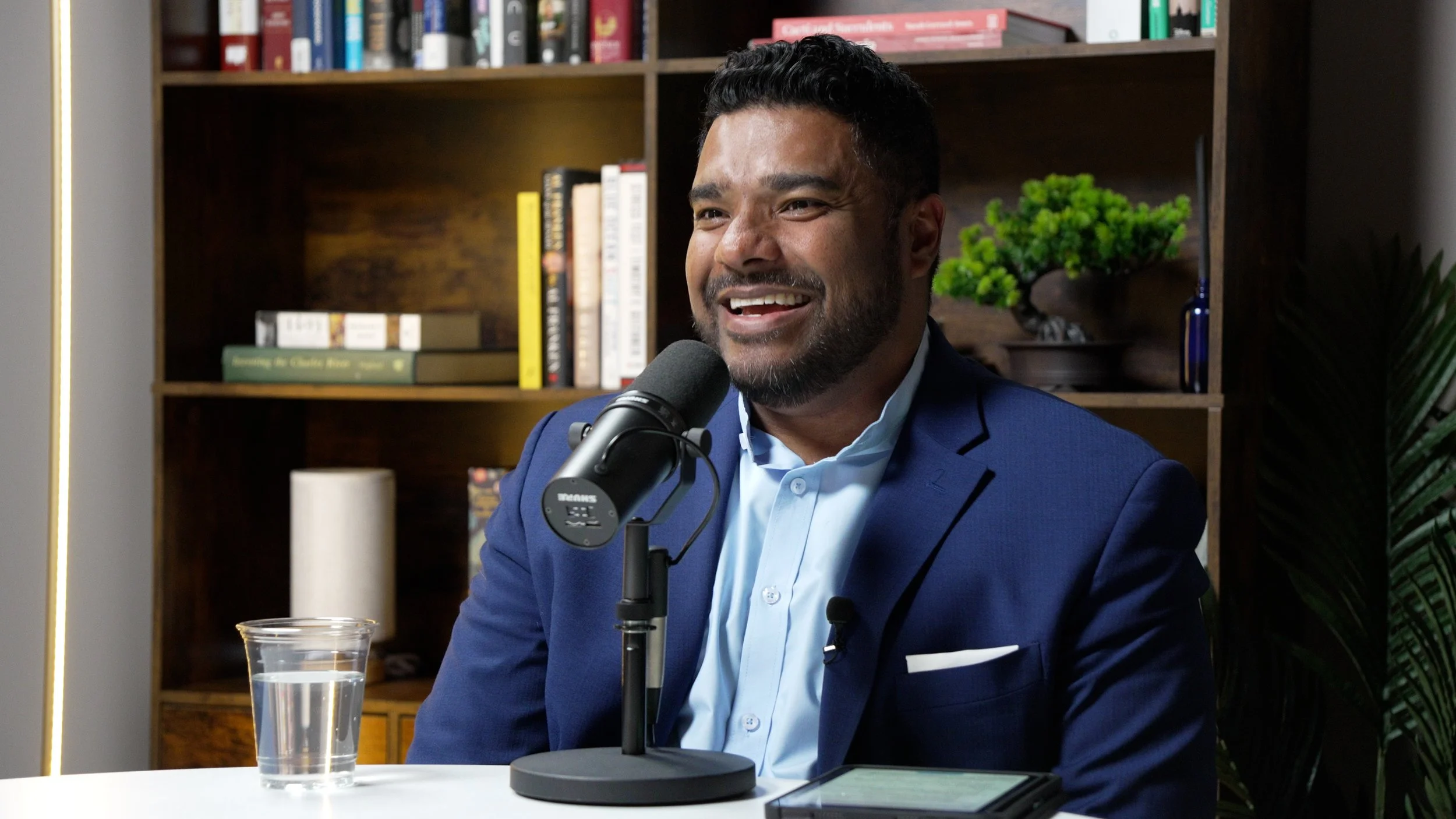 A man in a blue suit and light blue shirt speaking into a microphone during a podcast or interview, with a shelf of books and decorative plants in the background.