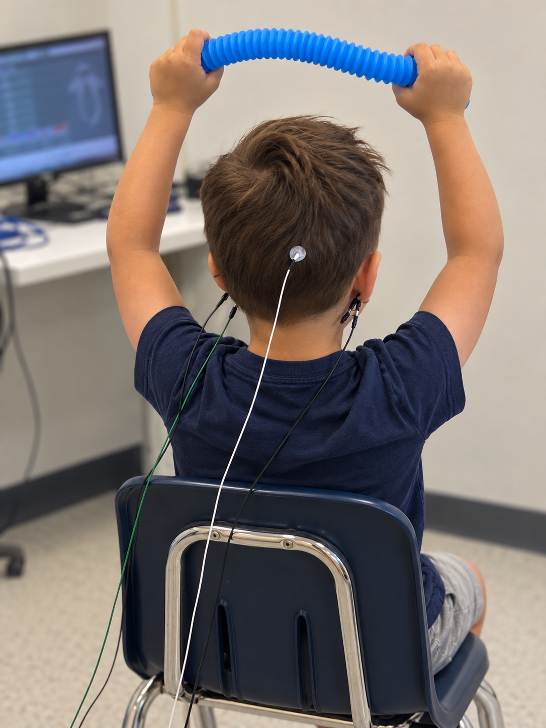 Child sitting in a chair with LENS  attached to his back, holding a blue flexible tube above his head, in a clinical setting with a computer monitor in the background.