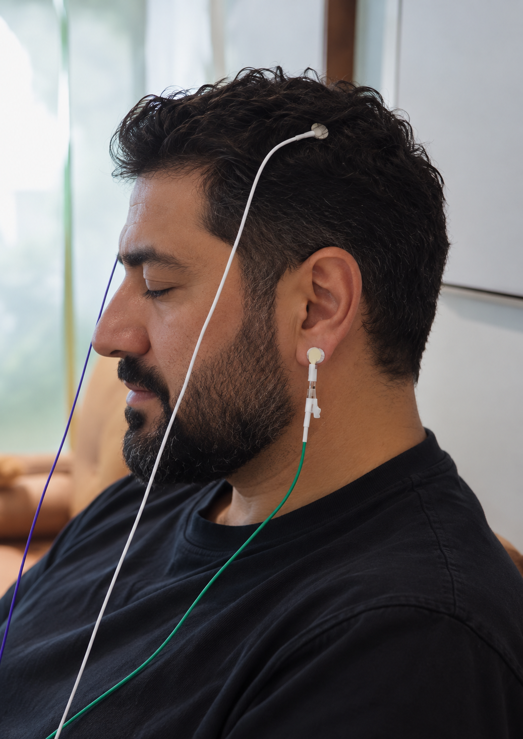 A man with closed eyes, wearing LENS monitors and a black shirt, is sitting with a calm expression.