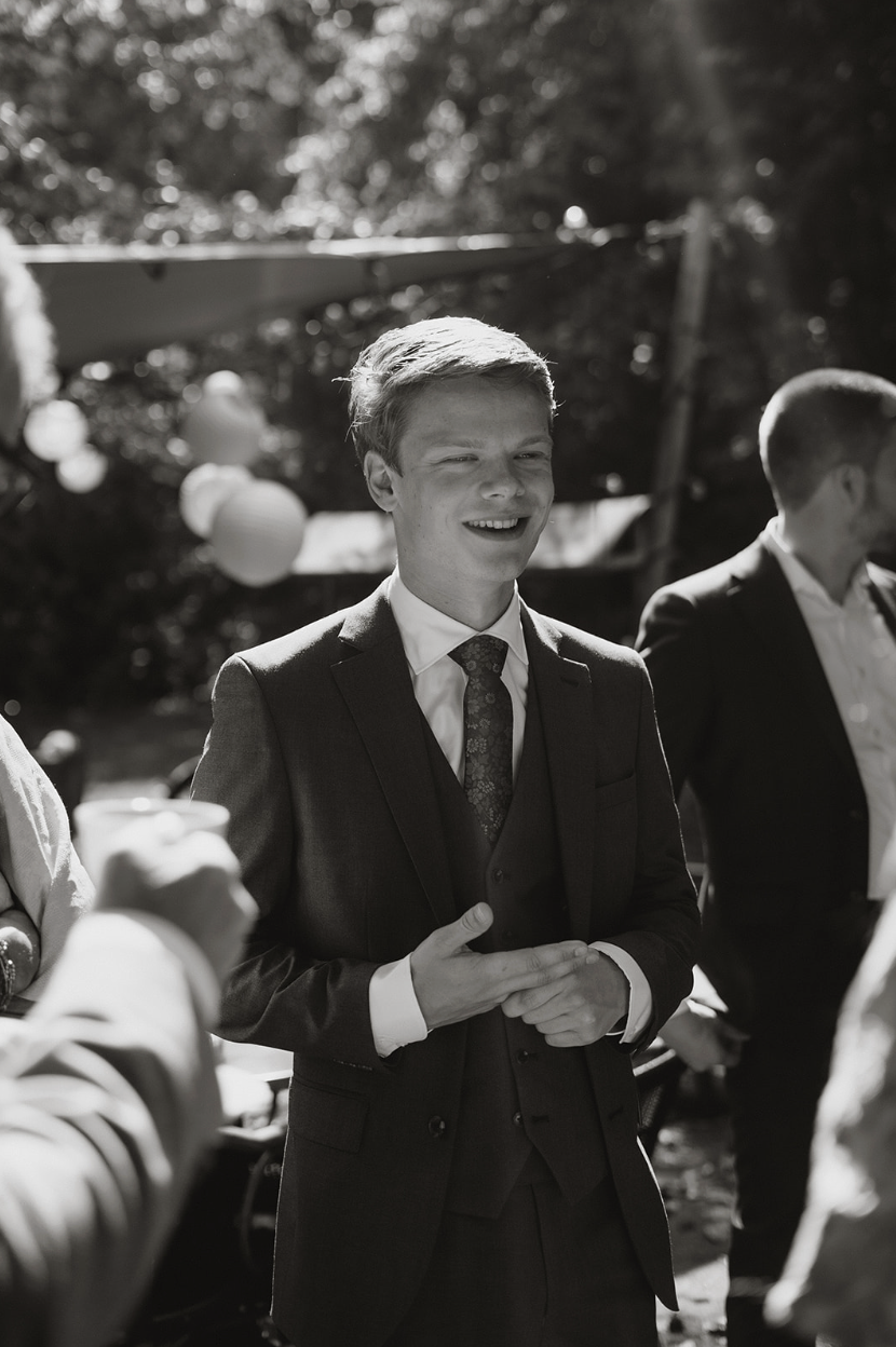 Een jonge man in een pak en stropdas, glimlachend tijdens een buitengelegen evenement, mogelijk een huwelijk of feest, met een houten structuur en papierlampjes op de achtergrond.