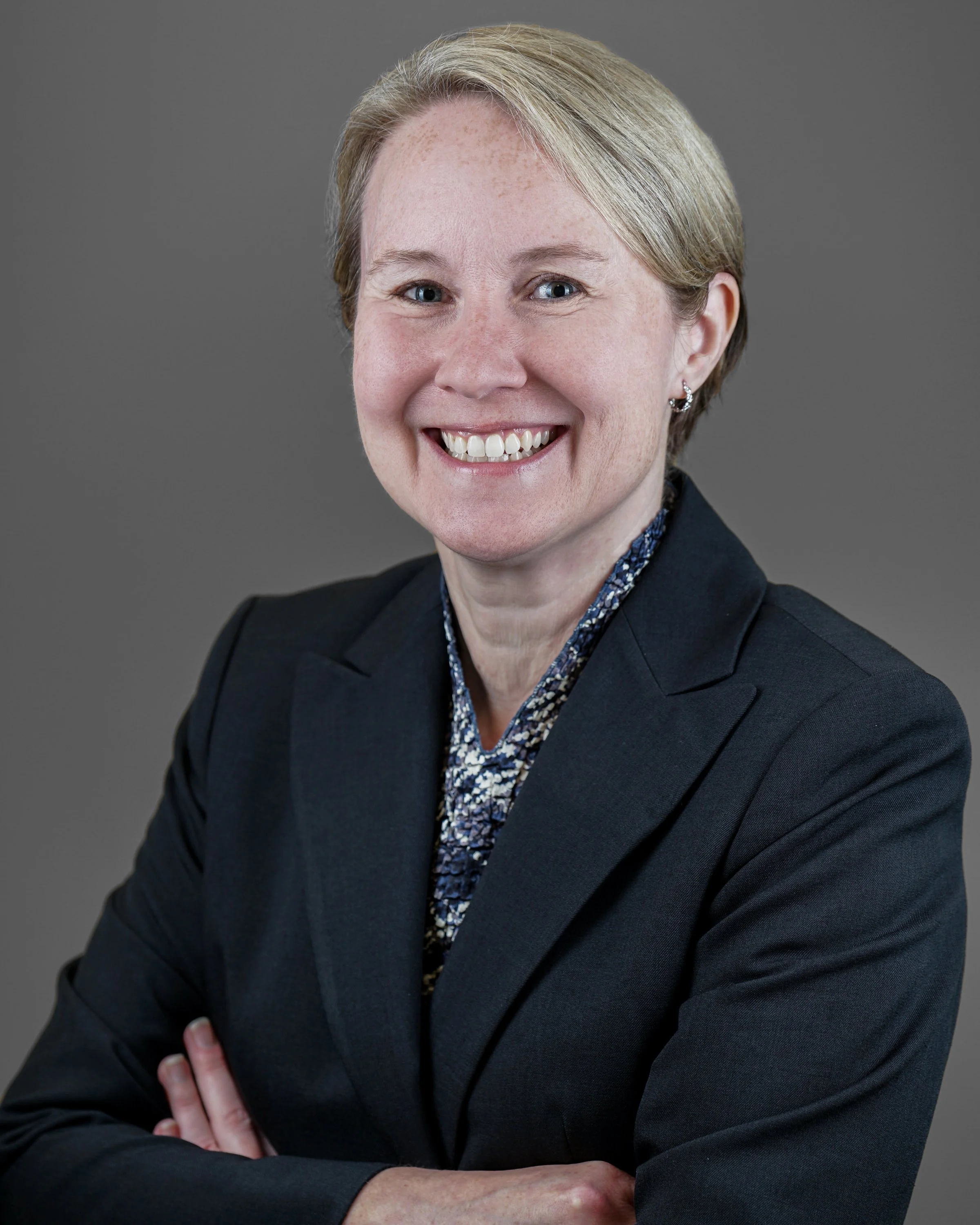 A woman with short blonde hair wearing a dark blazer and patterned blouse, smiling, with her arms crossed in front of a gray background.