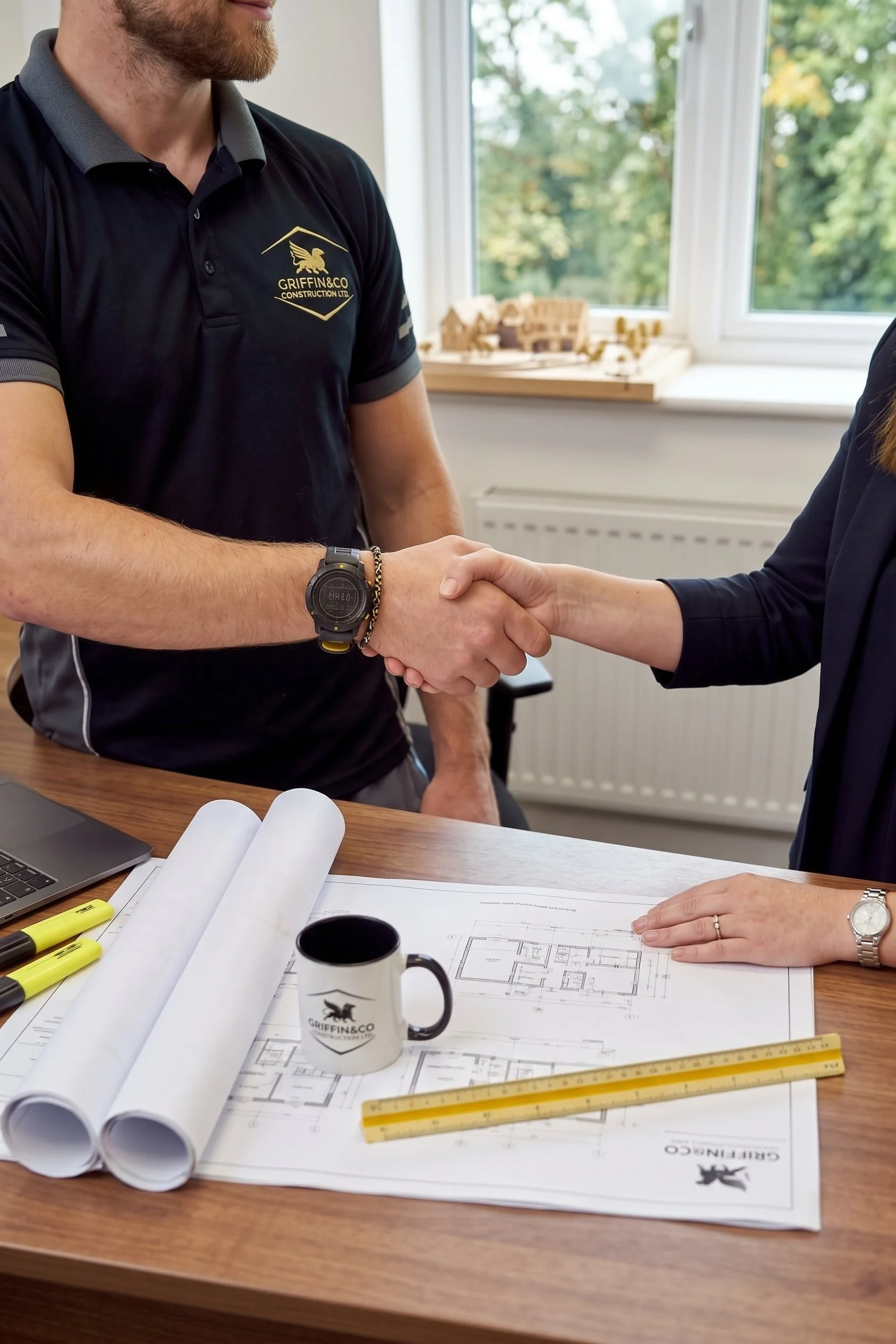 Handshake between two professionals at a work desk with blueprints, architectural plans, highlighters, coffee mug, rulers, and a laptop in a bright office.