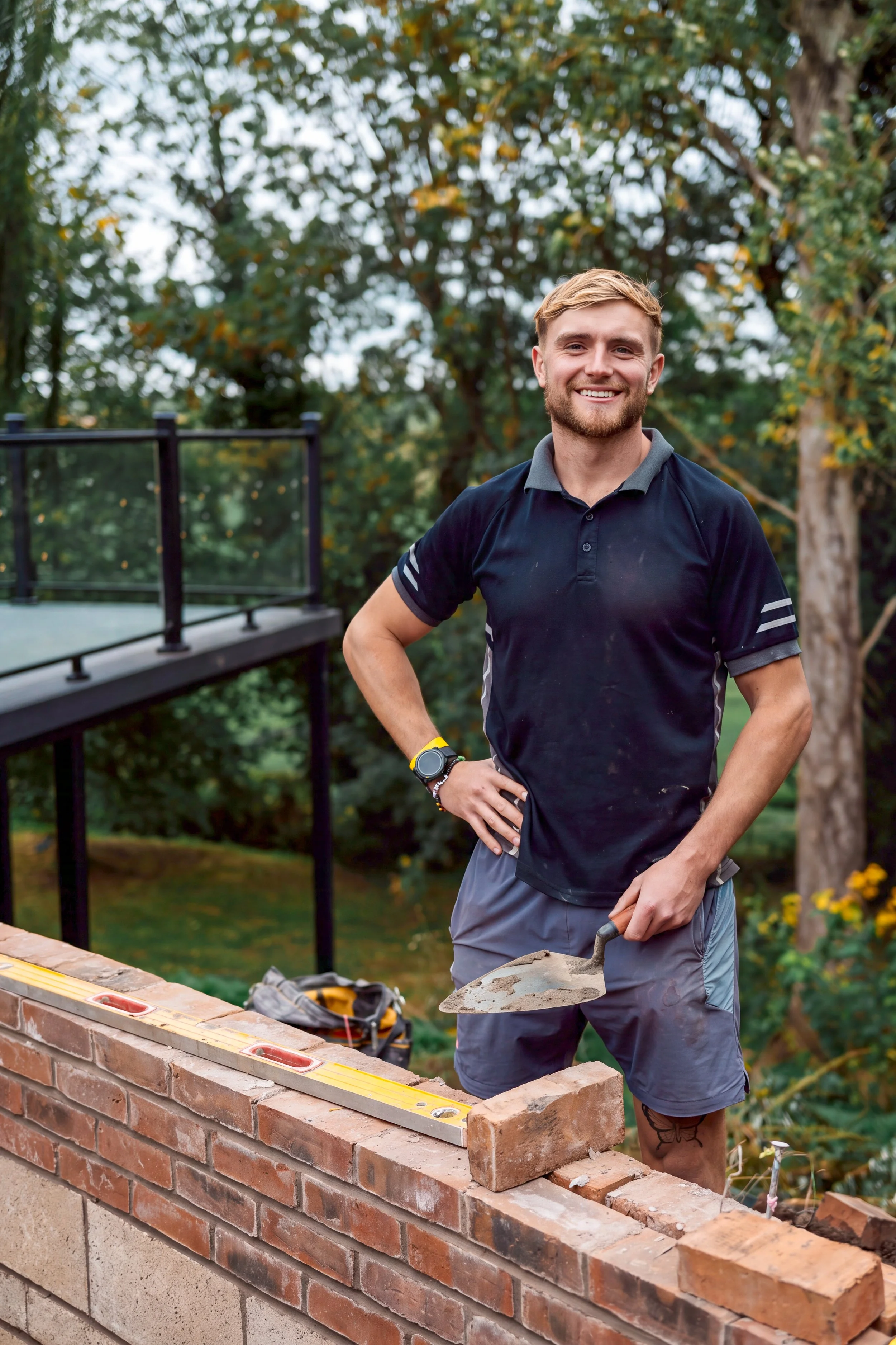 A young man with a beard and tattoos, wearing a black athletic shirt and gray shorts, is building a brick wall outdoors. He is smiling and holding a trowel, with construction tools and a spirit level on the wall in front of him. There are trees and greenery in the background.