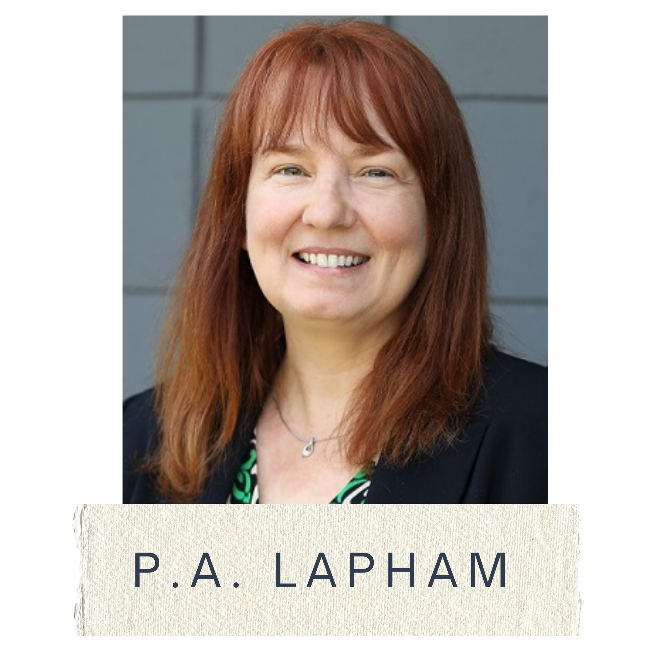 Close-up of a woman with red hair smiling, standing in front of a gray wall, at an event with a sign that reads 'P.A. Lapham' in the foreground.
