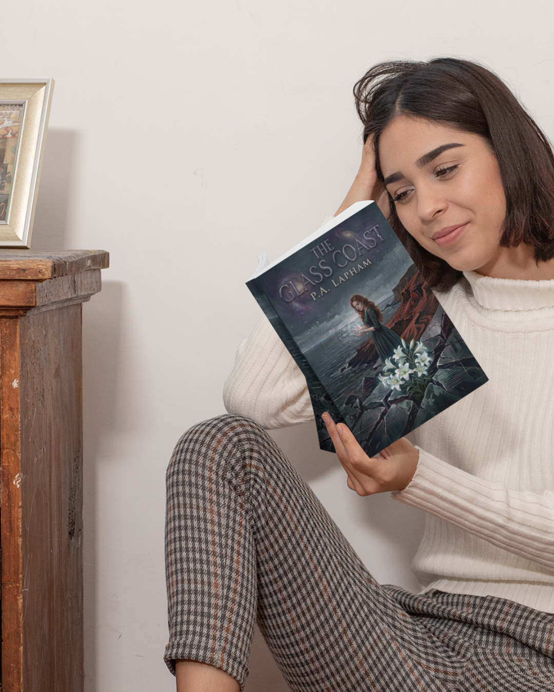 A woman sitting on the floor, leaning against a white wall, holding and reading a book titled 'The Glass Coast' by P. A. Lapham. She has short brown hair, is wearing a white knitted sweater, and plaid pants. There is a small wooden table with a framed photo on top next to her.