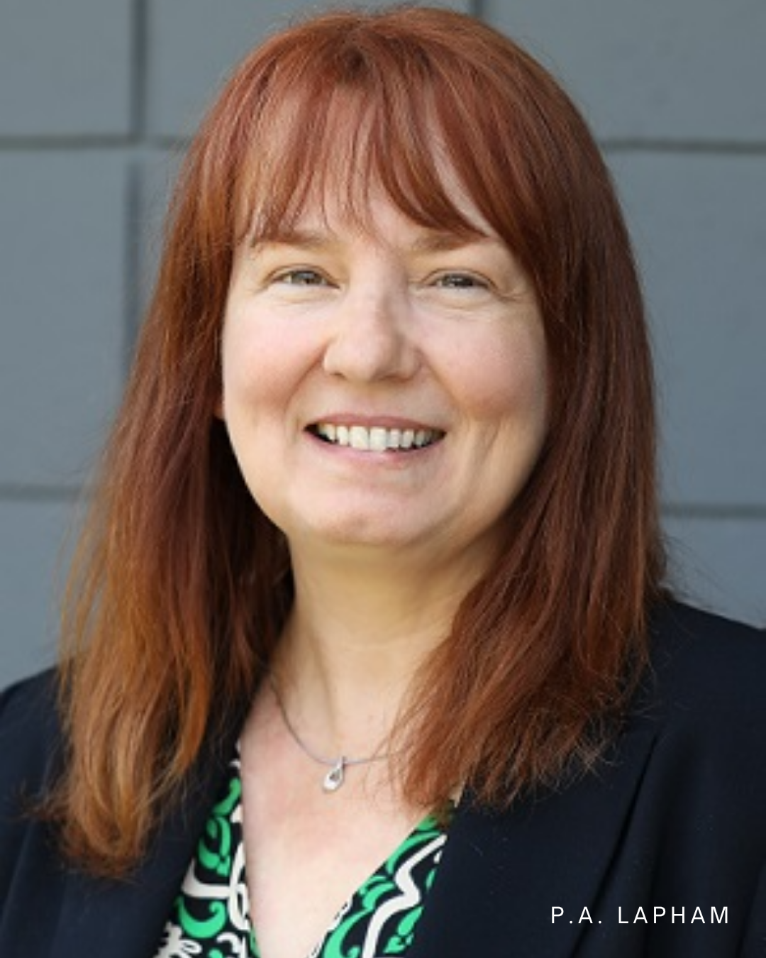 Portrait of a smiling woman with long reddish-brown hair, wearing a black blazer and a patterned top, standing against a gray wall.