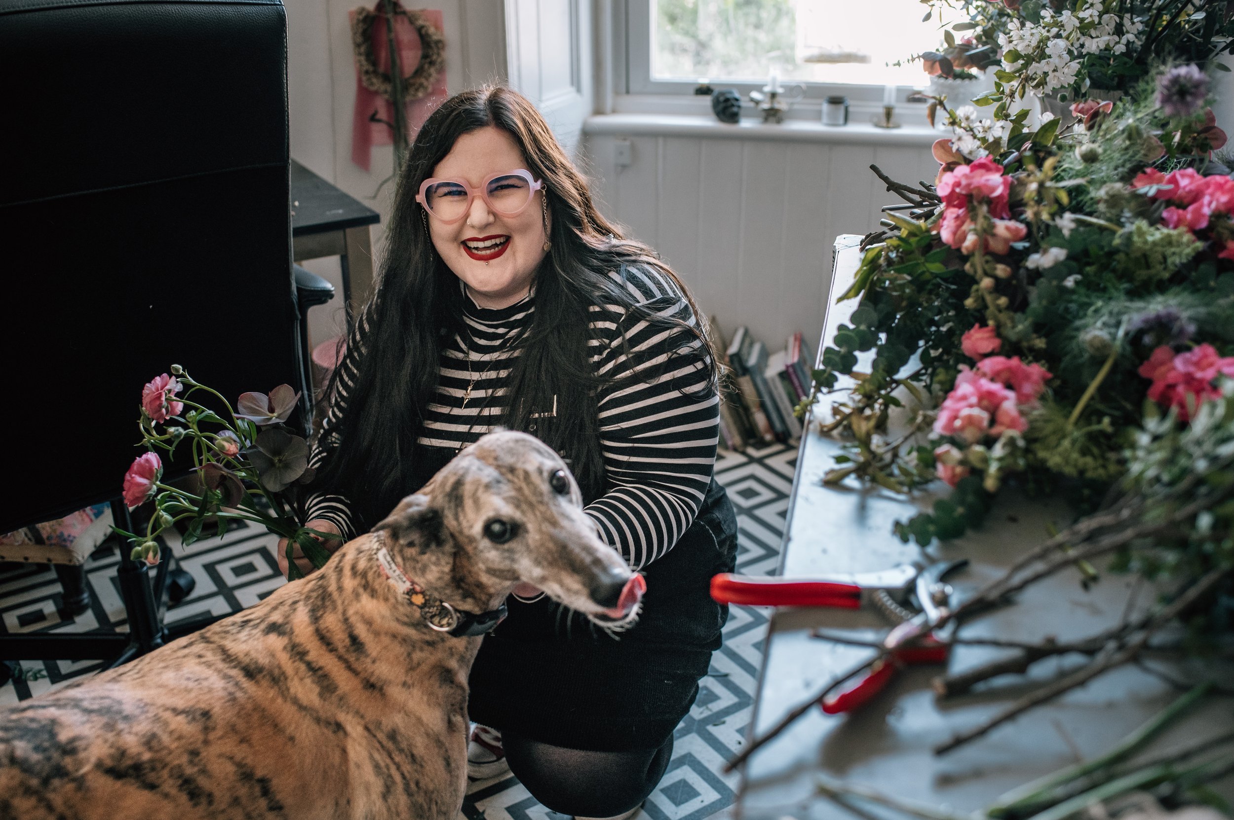 Sarah Rushbrooke has long dark hair, is wearing glasses, a striped shirt, and makeup, smiling while playing with a brindle-coloured dog in her Glasgow flower studio. There are flowers in the foreground, a window with white trim & floral arrangement